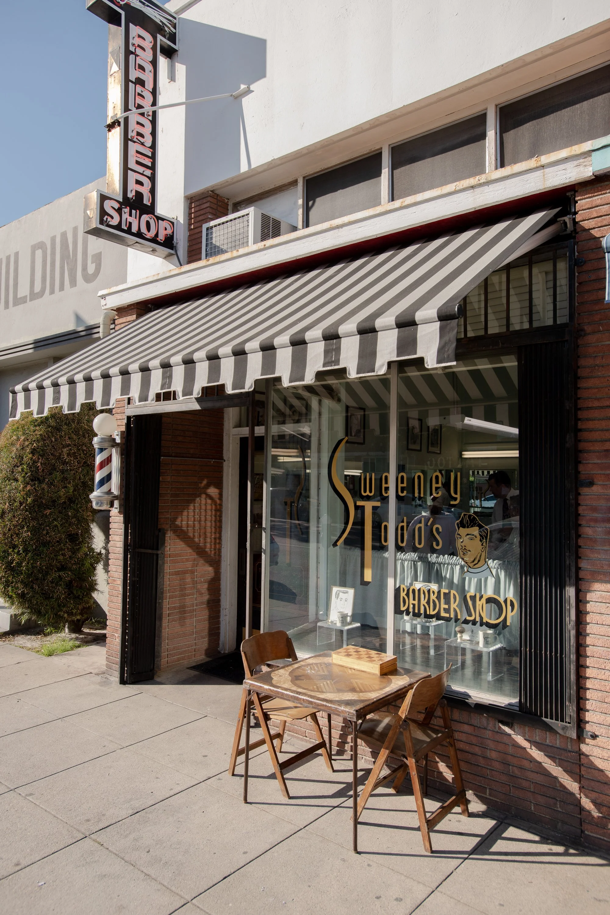 Exterior of Sweeney Todd's Barbershop in Los Angeles showing striped black and white awning, gold script window lettering, barber pole, and sidewalk table with chairs