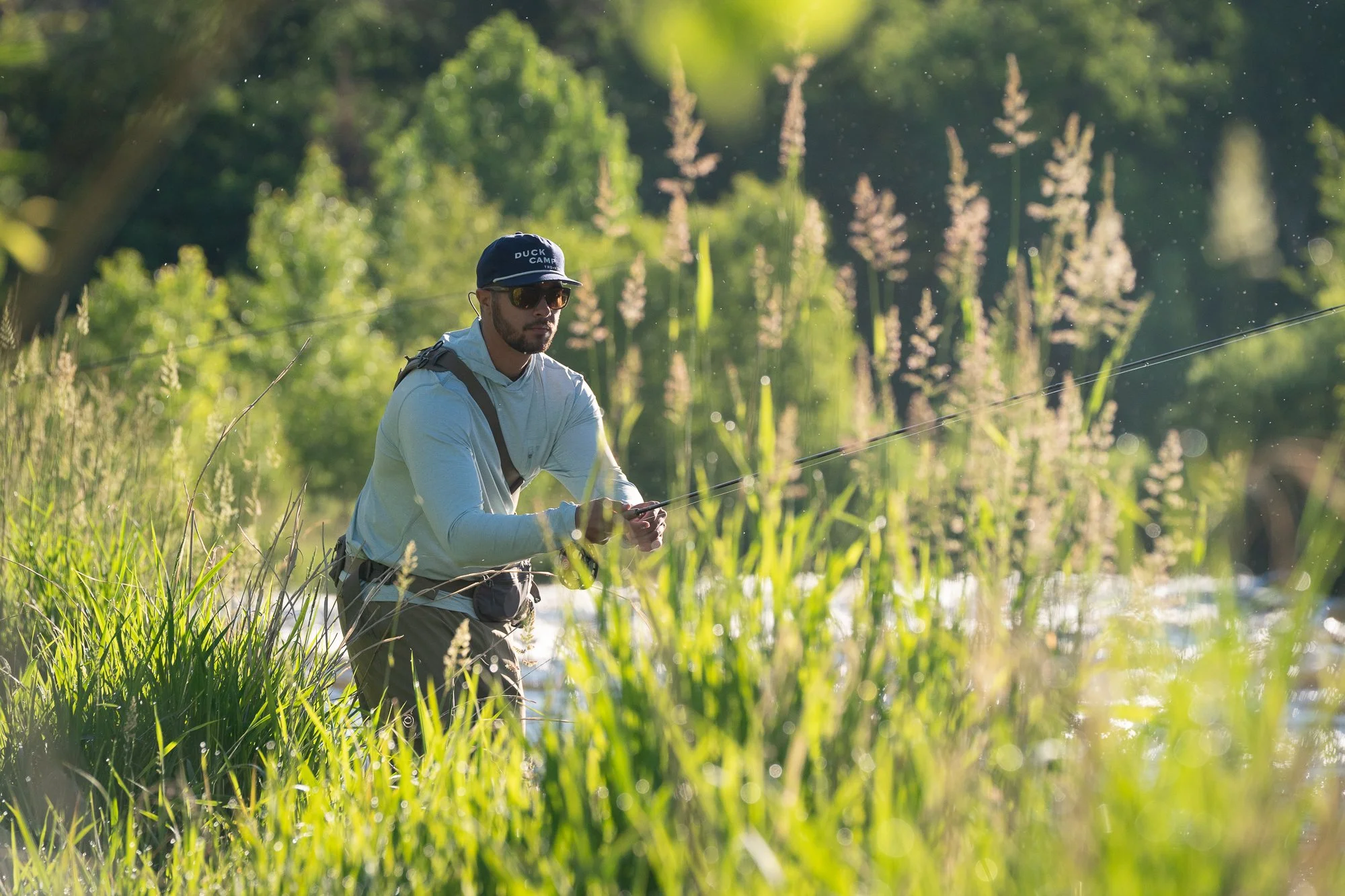 An angler wearing a Duck Camp hat works a fly rod while standing in tall grass and river current