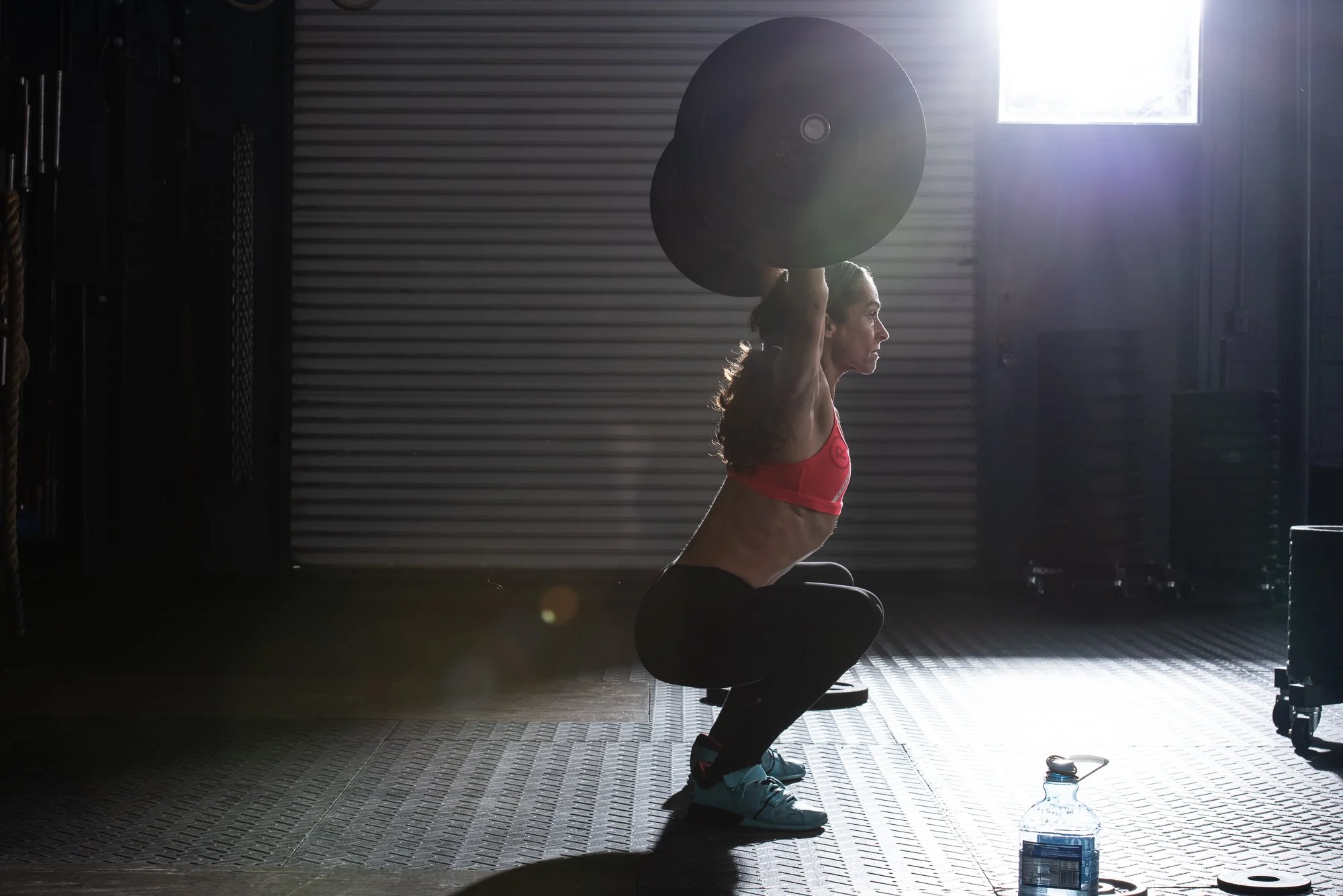 Female athlete performing overhead barbell squat in crossfit gym with strong backlighting and water bottle on floor