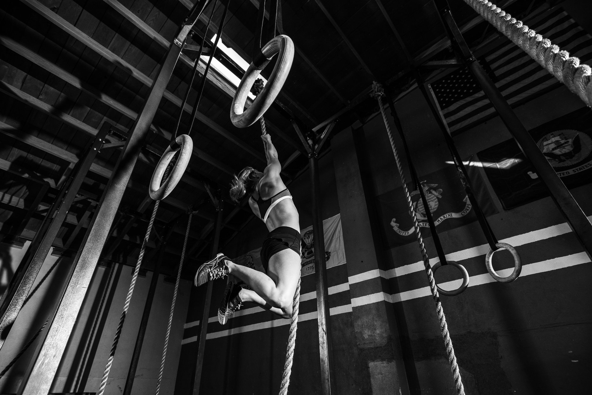 Female CrossFit athlete climbing a rope beneath gymnastic rings inside a gritty gym.