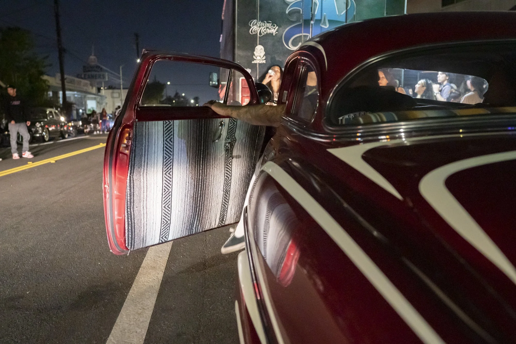 Open door of a deep maroon lowrider showing a serape-upholstered interior panel on a Long Beach street at night during the Syndicate Barbershop anniversary