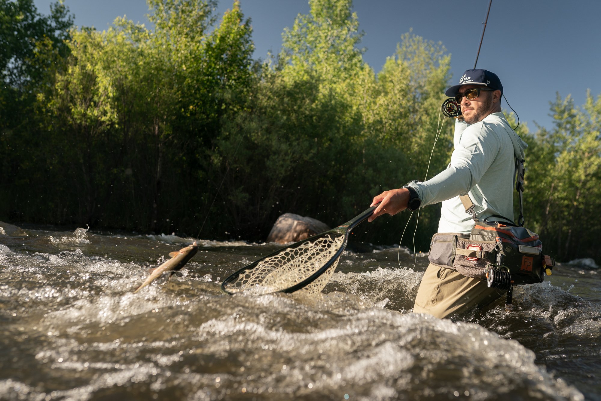 An angler reaches a landing net toward a fish while standing in fast river current