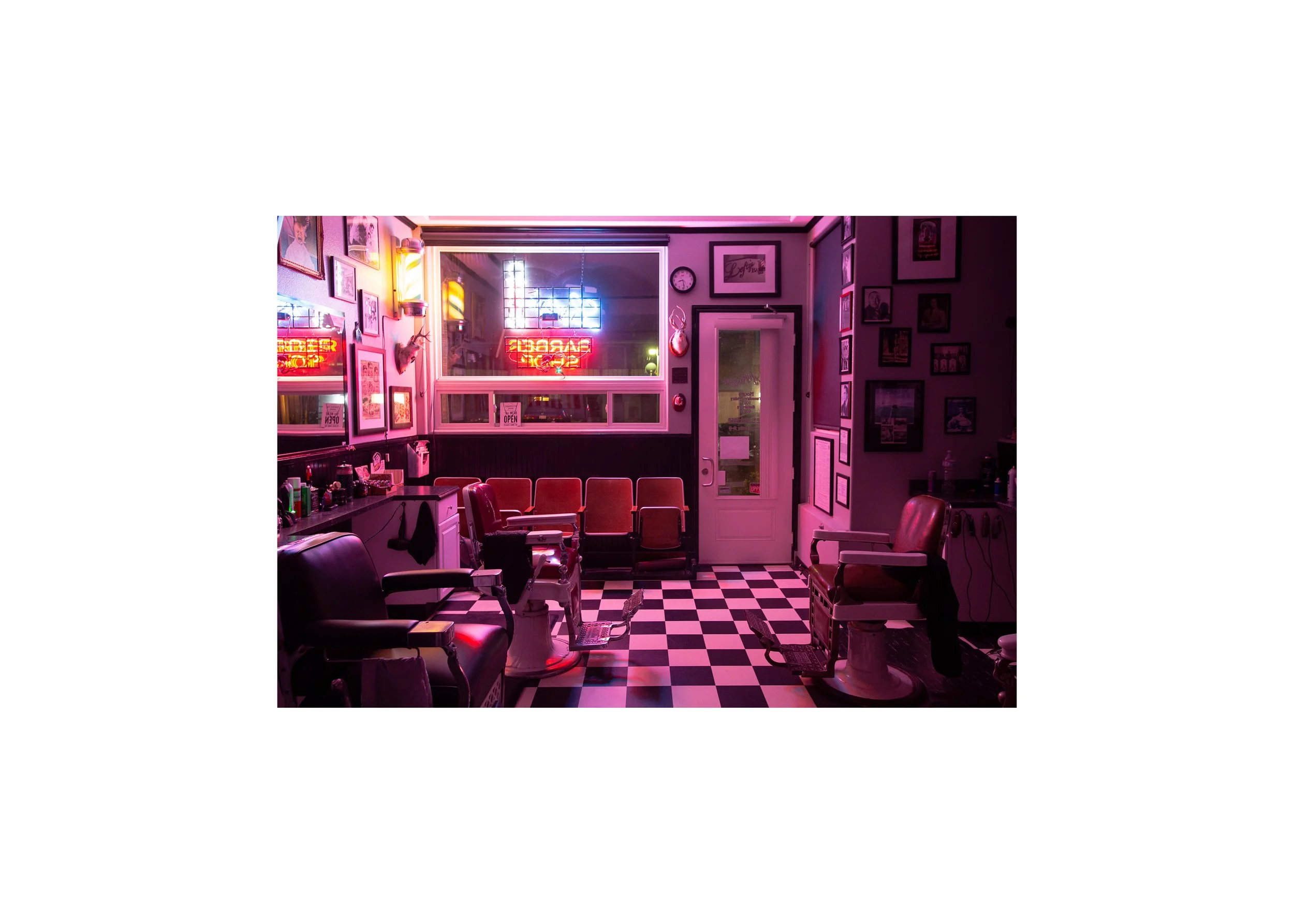 Interior of a vintage barbershop at night with empty barber chairs, pink neon sign, checkered floor, and framed photos on the walls.