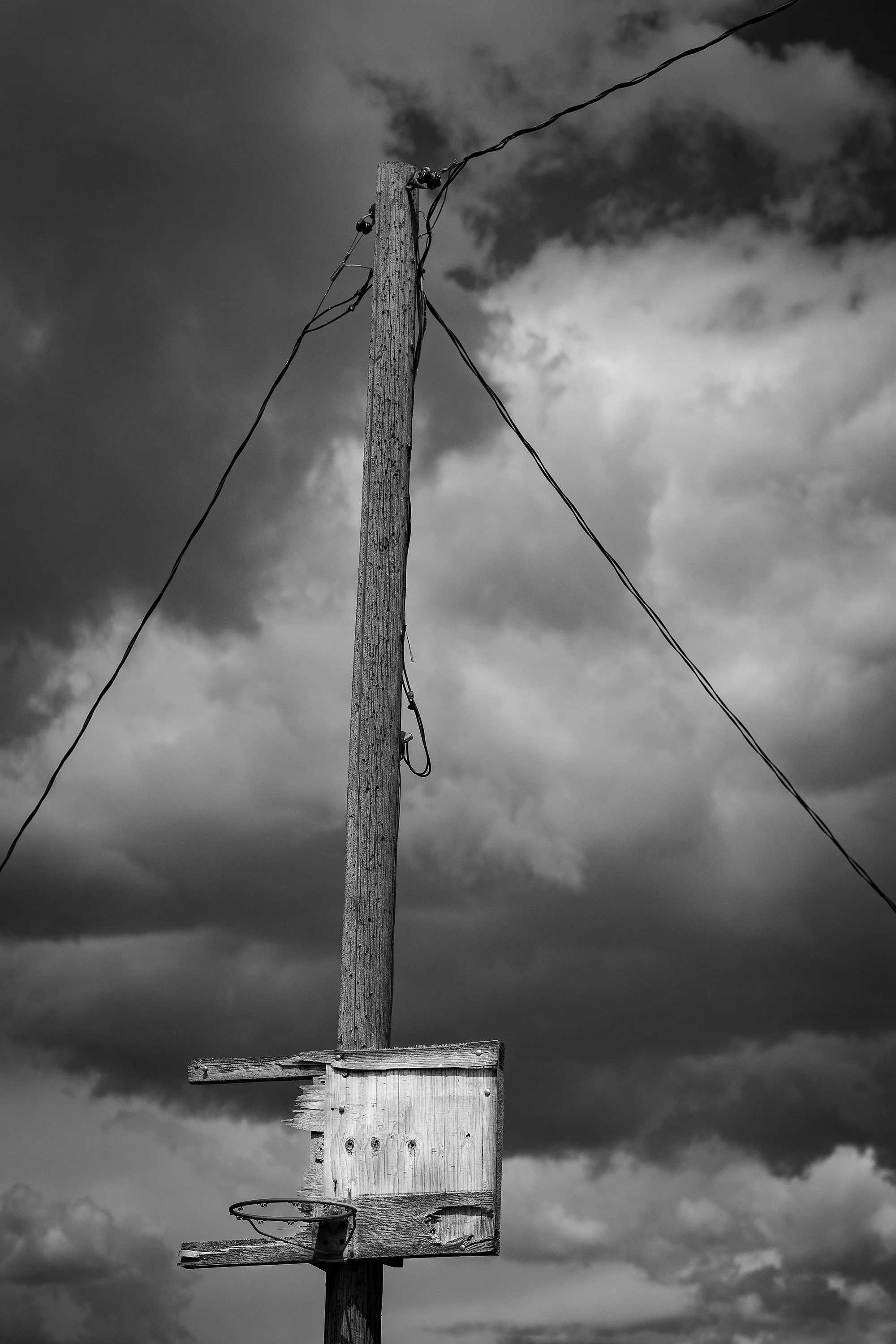 Weathered wooden basketball backboard and hoop mounted to a utility pole beneath a cloudy sky.