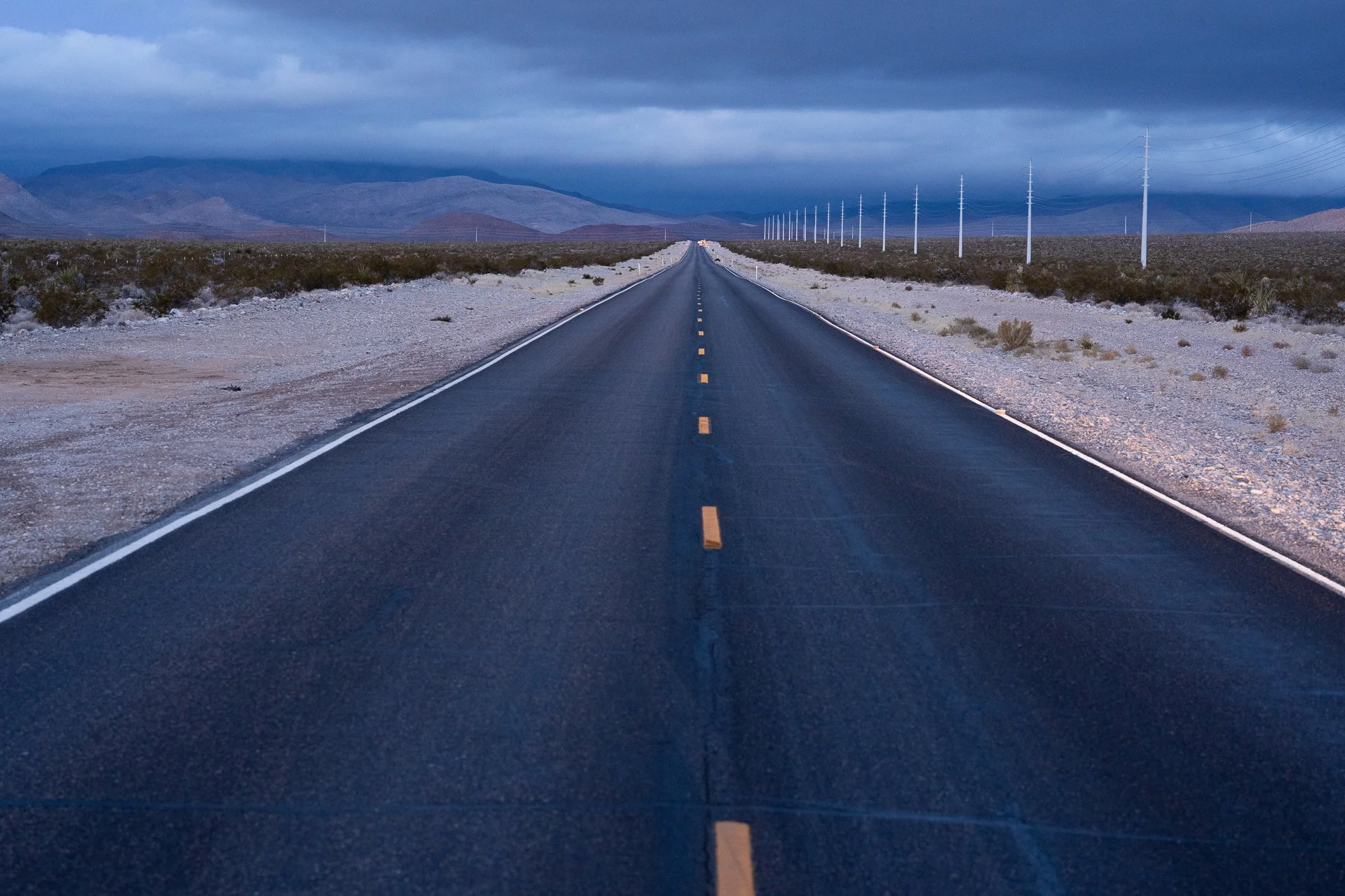 Straight two-lane Nevada highway at dusk with utility poles receding into the distance, dark storm clouds overhead, and mountains visible on the horizon.