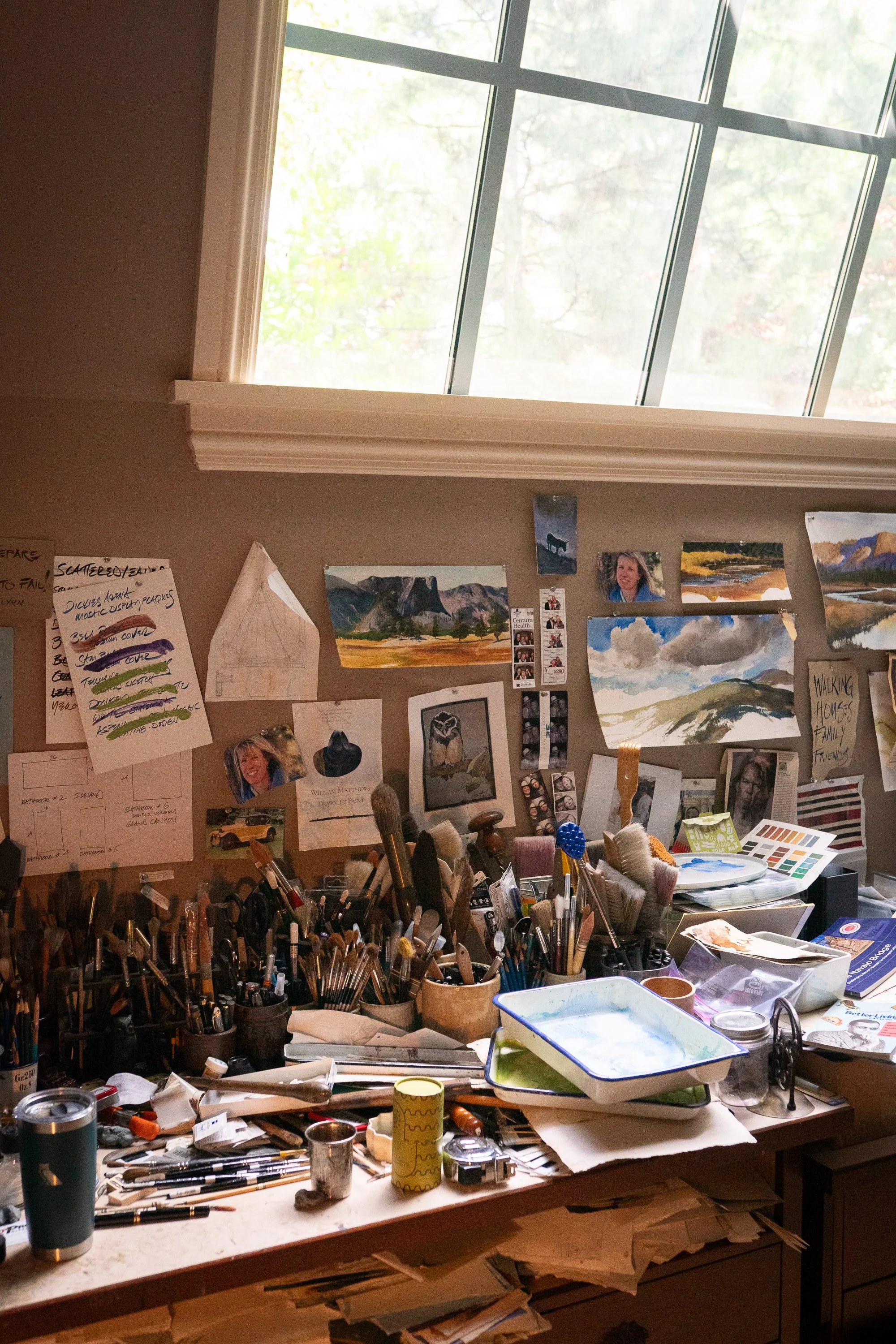 Artist desk filled with brushes, pinned sketches, reference photos and watercolor trays in William Matthews’ studio