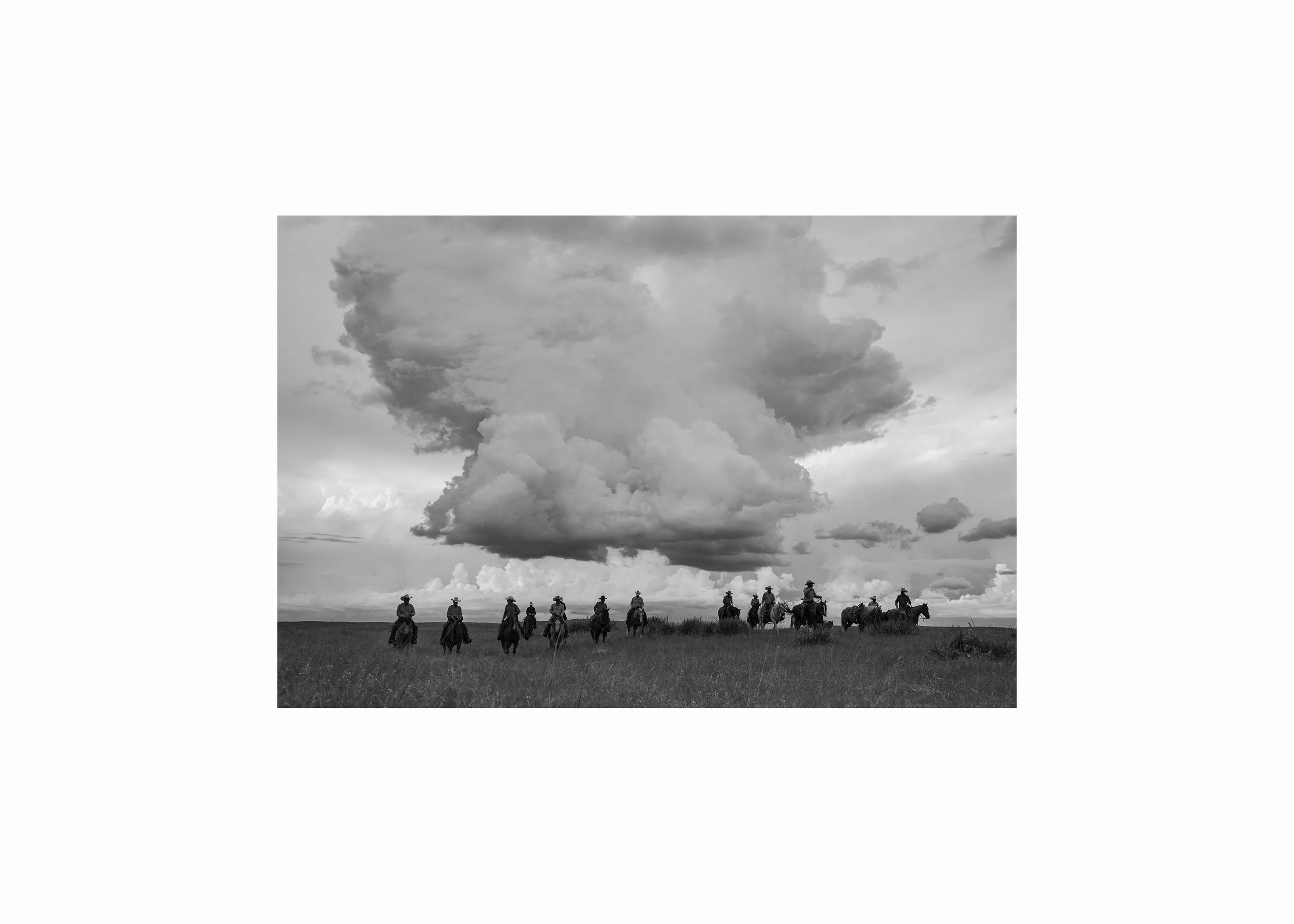 Black and white photograph of cowboys on horseback riding through open country beneath large storm clouds, photographed by Rob Hammer.