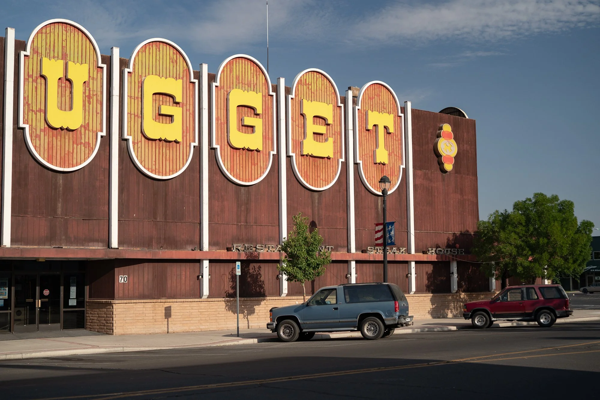 Large Nugget Casino restaurant and steakhouse exterior with oversized yellow letter signage on a brown facade, two SUVs parked out front, street trees along the sidewalk.