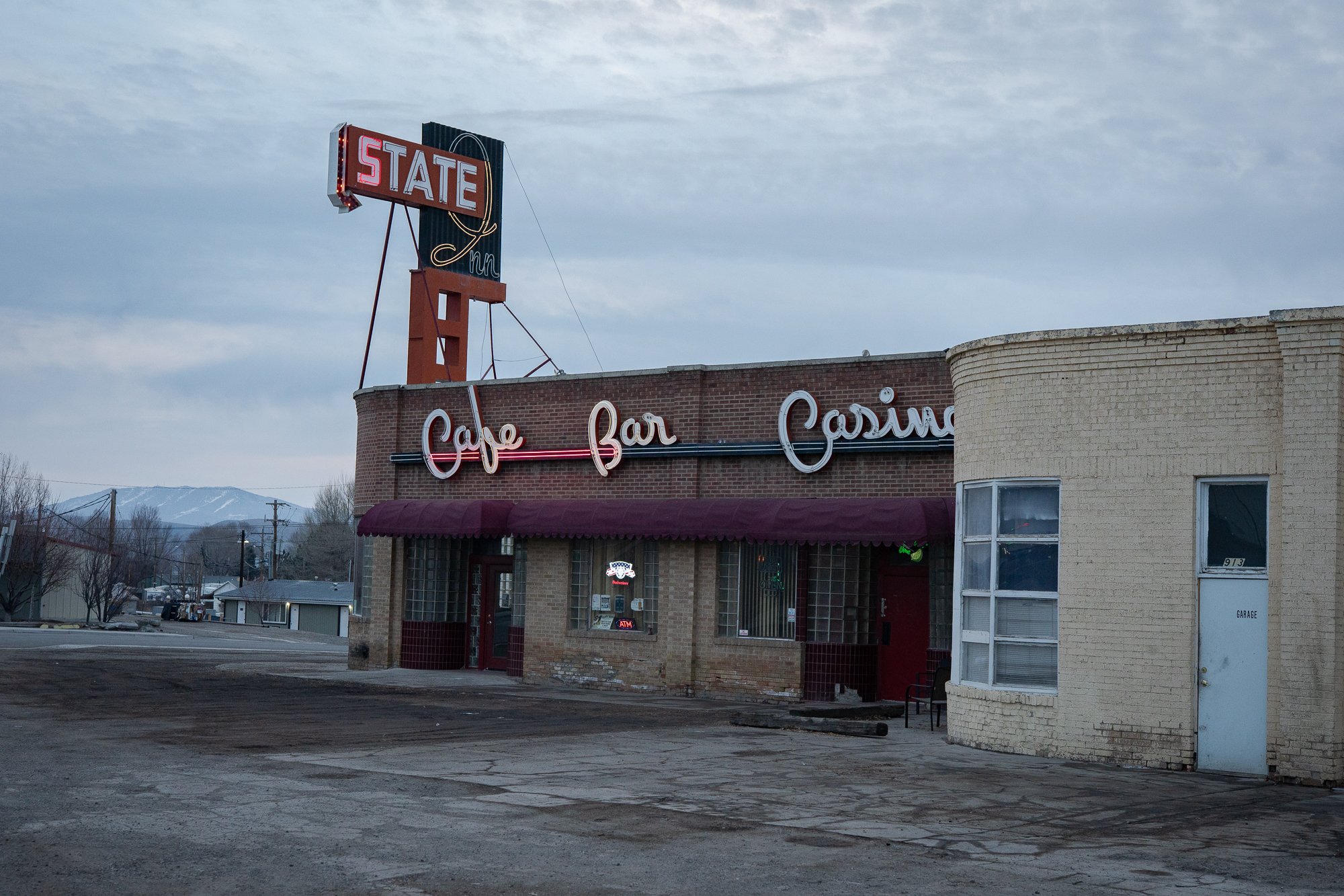 Brick Nevada roadside building with a lit State Inn neon sign and Cafe Bar Casino facade lettering, empty gravel lot, overcast sky.