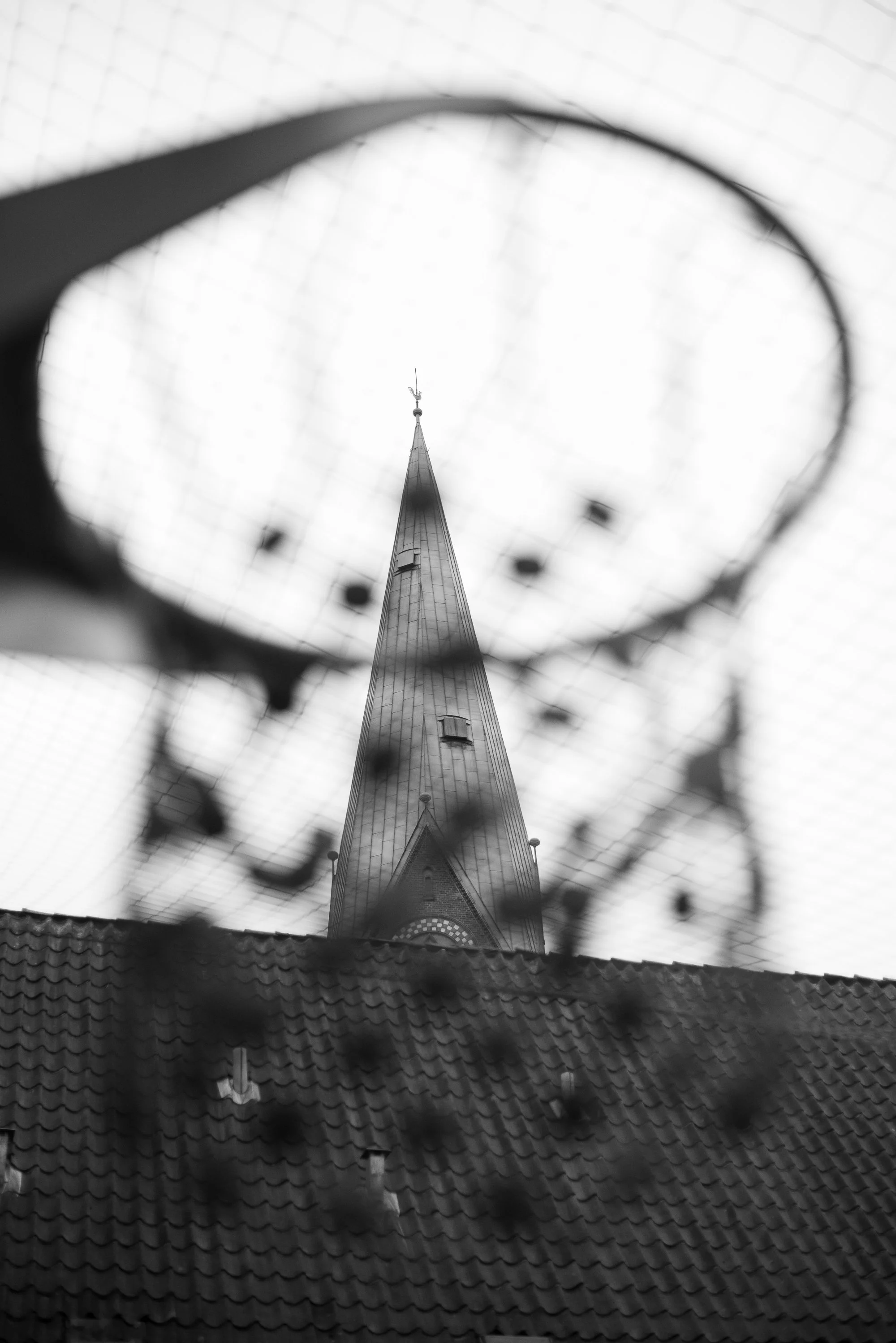 Black and white photograph of a church steeple framed through a basketball hoop on a European outdoor court.