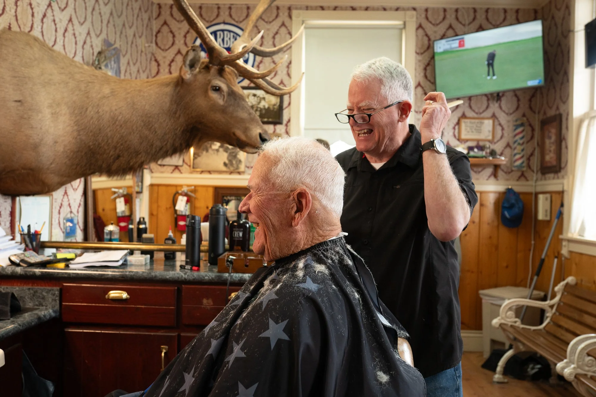 Old school barbershop haircut with a barber laughing beside a client seated beneath a mounted elk head.