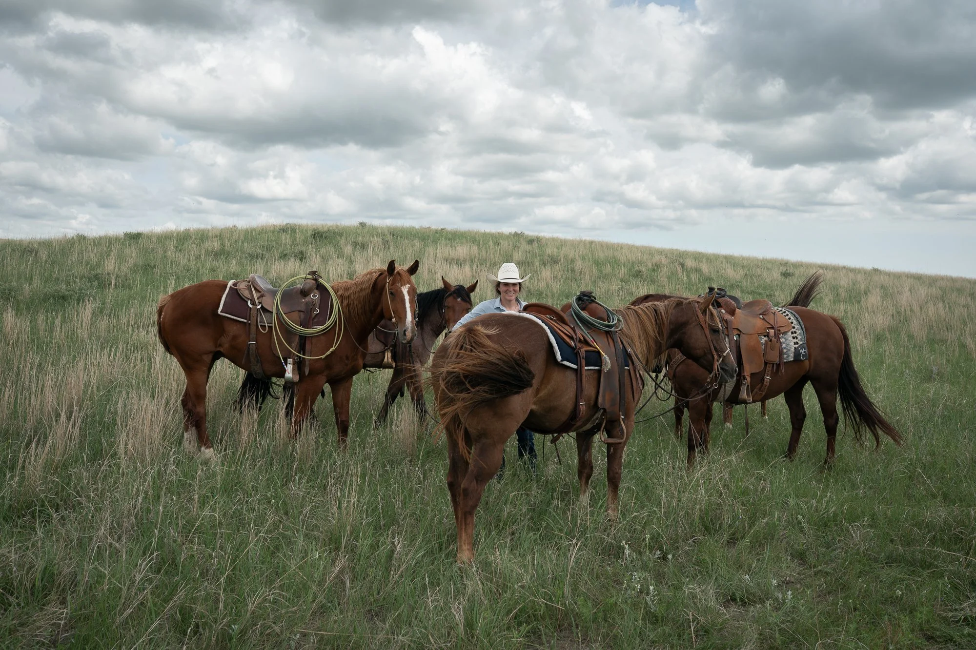 Cowgirl standing among saddled ranch horses in tall prairie grass at Haythorn Ranch in Nebraska.
