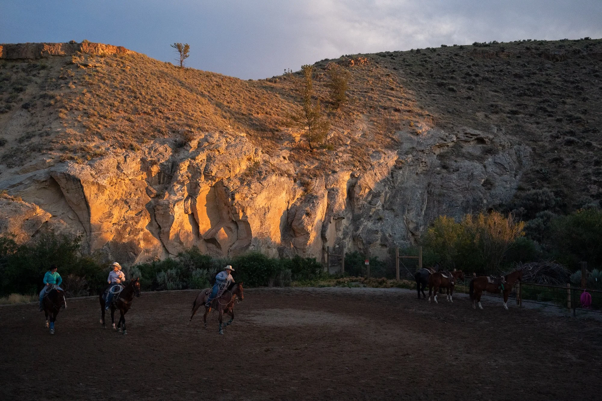 Cowboys on horseback prepare near the arena beneath sunlit cliffs before the Dubois Rodeo in Wyoming
