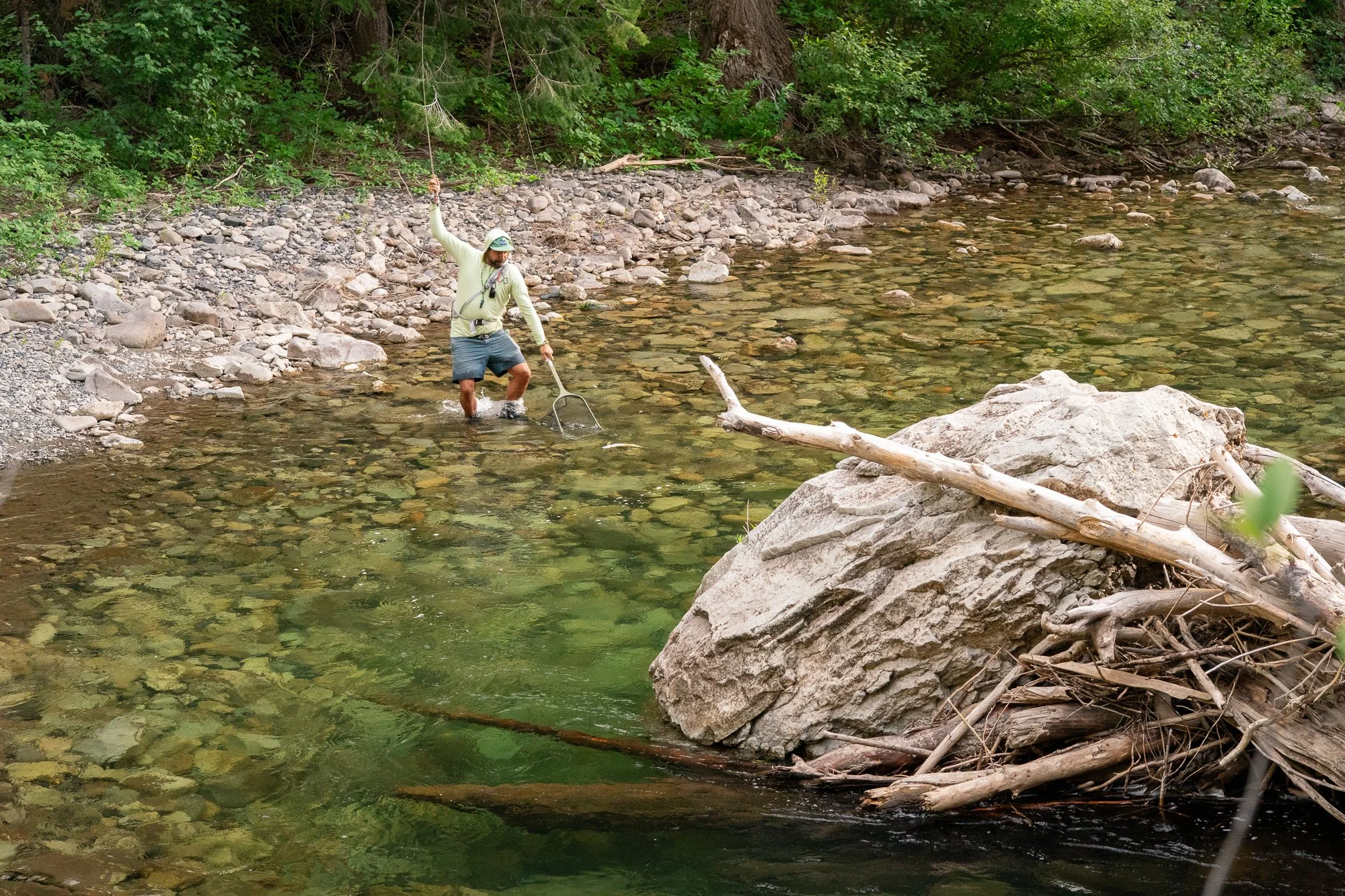 A fly angler stands in clear river water with a landing net raised beside him