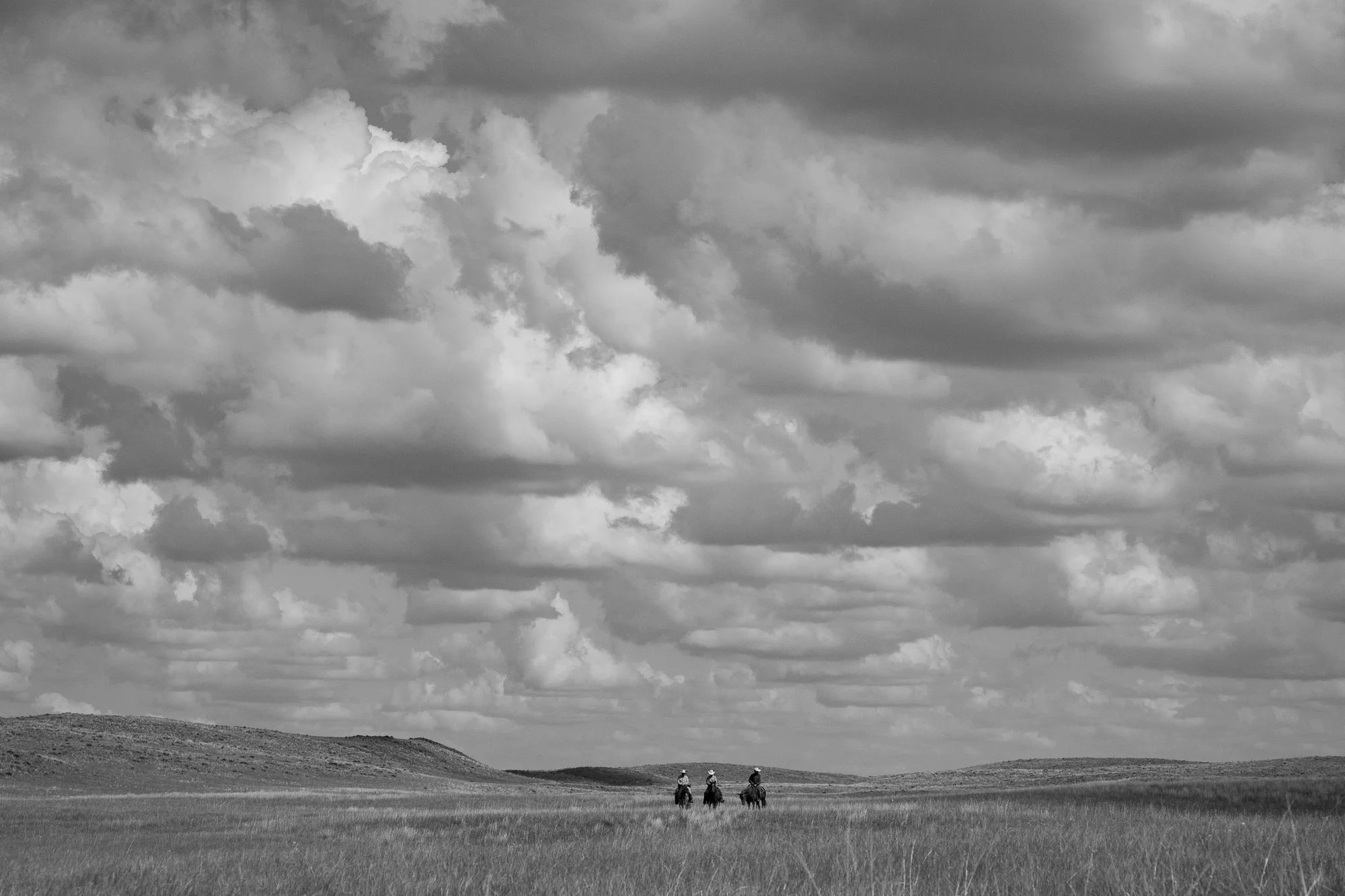 Three cowboys ride across the open prairie of the Nebraska Sandhills at Haythorn Ranch beneath dramatic summer clouds.