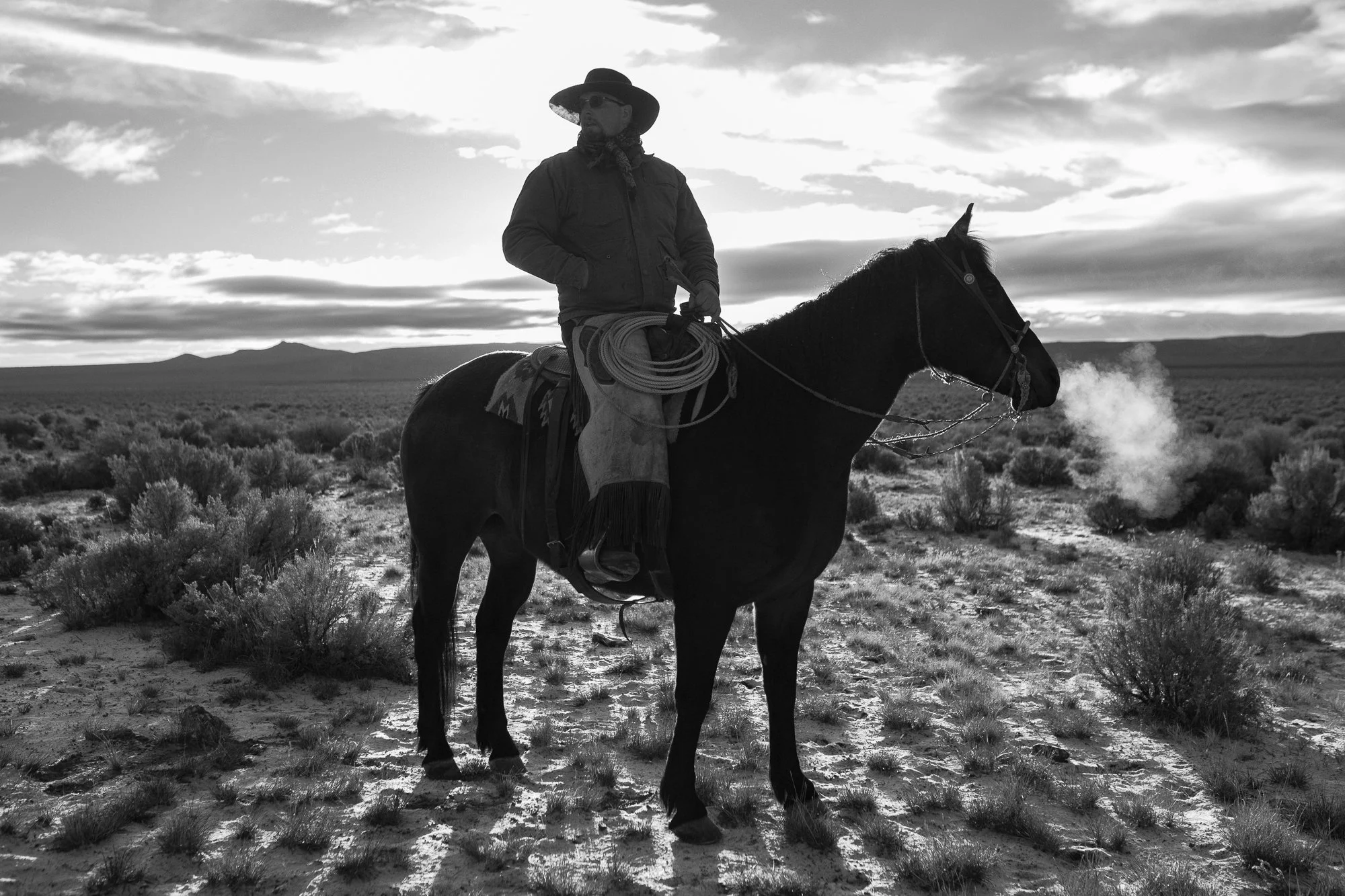 A cowboy sits on horseback in black and white with open high desert country behind him