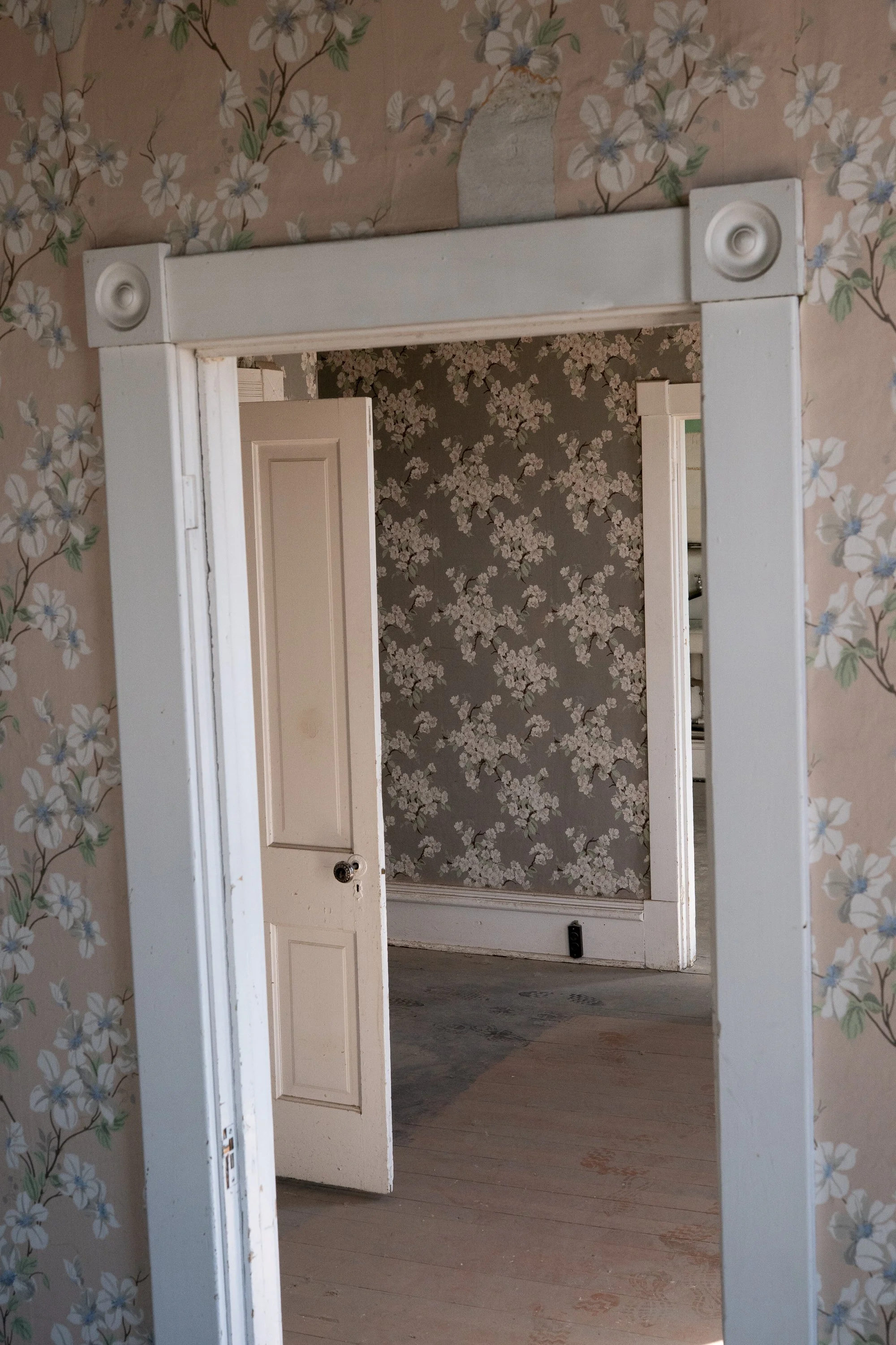 Open white doorway framed by faded floral wallpaper, with a second doorway and worn wooden flooring in an older interior in Douglas, Wyoming.