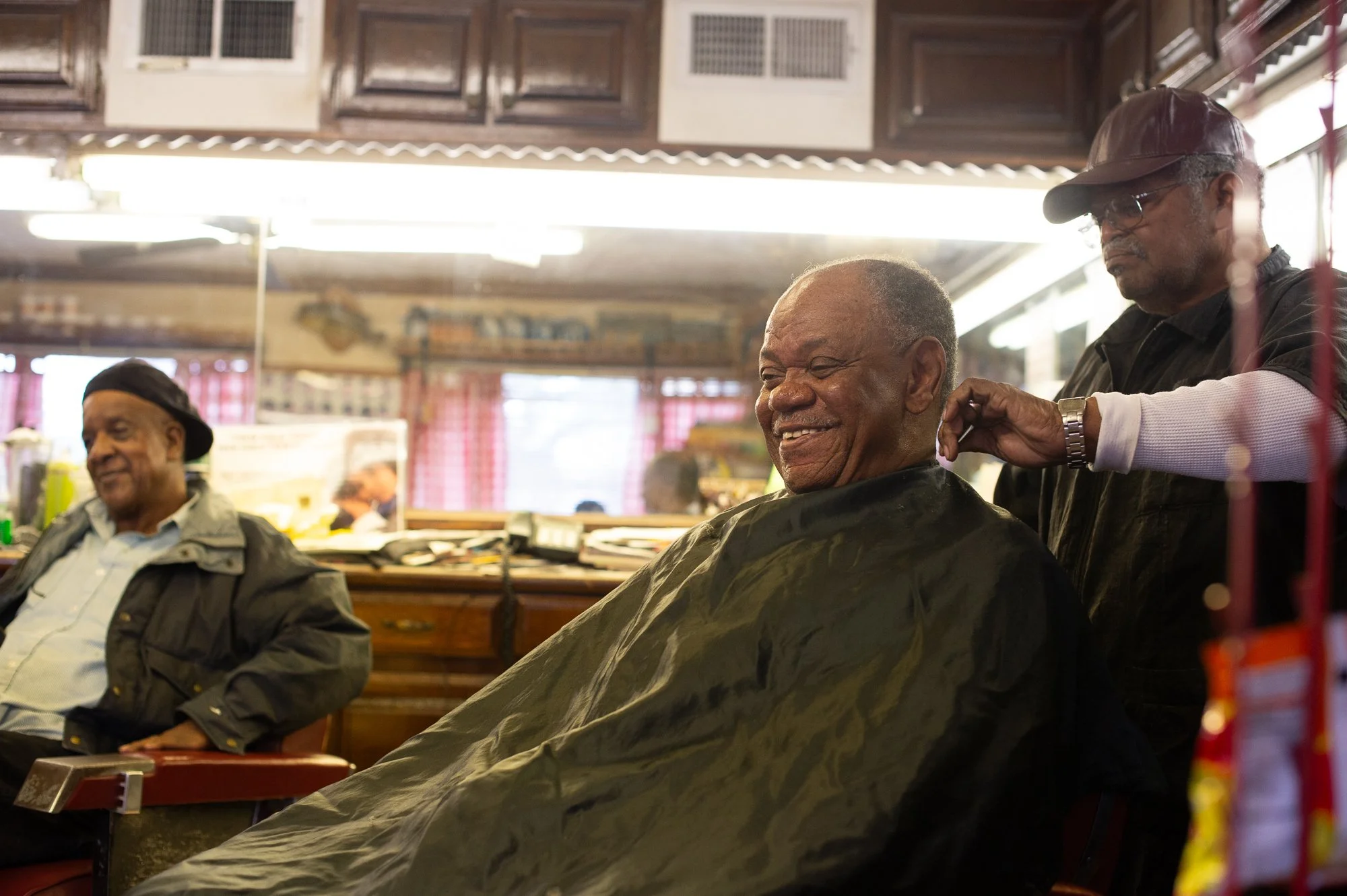 Barber trimming a client’s hair with another man sitting nearby in a classic barbershop interior
