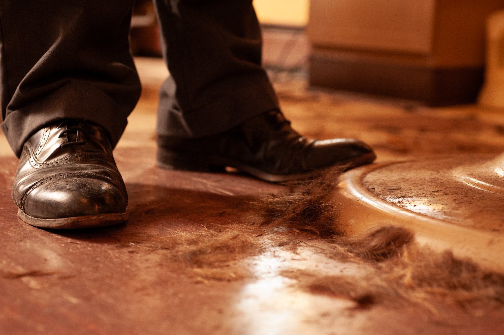 Hair clippings on the floor beside a barber's shoes and chair at Pugsly's SideShow Barbershop.