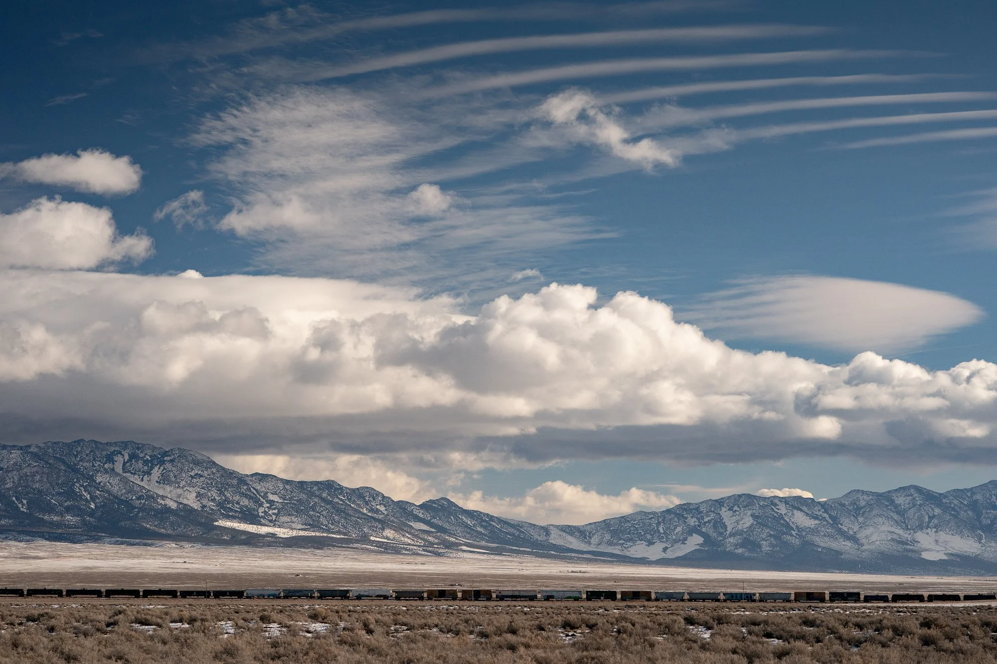 Long freight train crossing a flat Nevada desert basin with snow-dusted mountains and large layered cloud formations filling most of the sky.