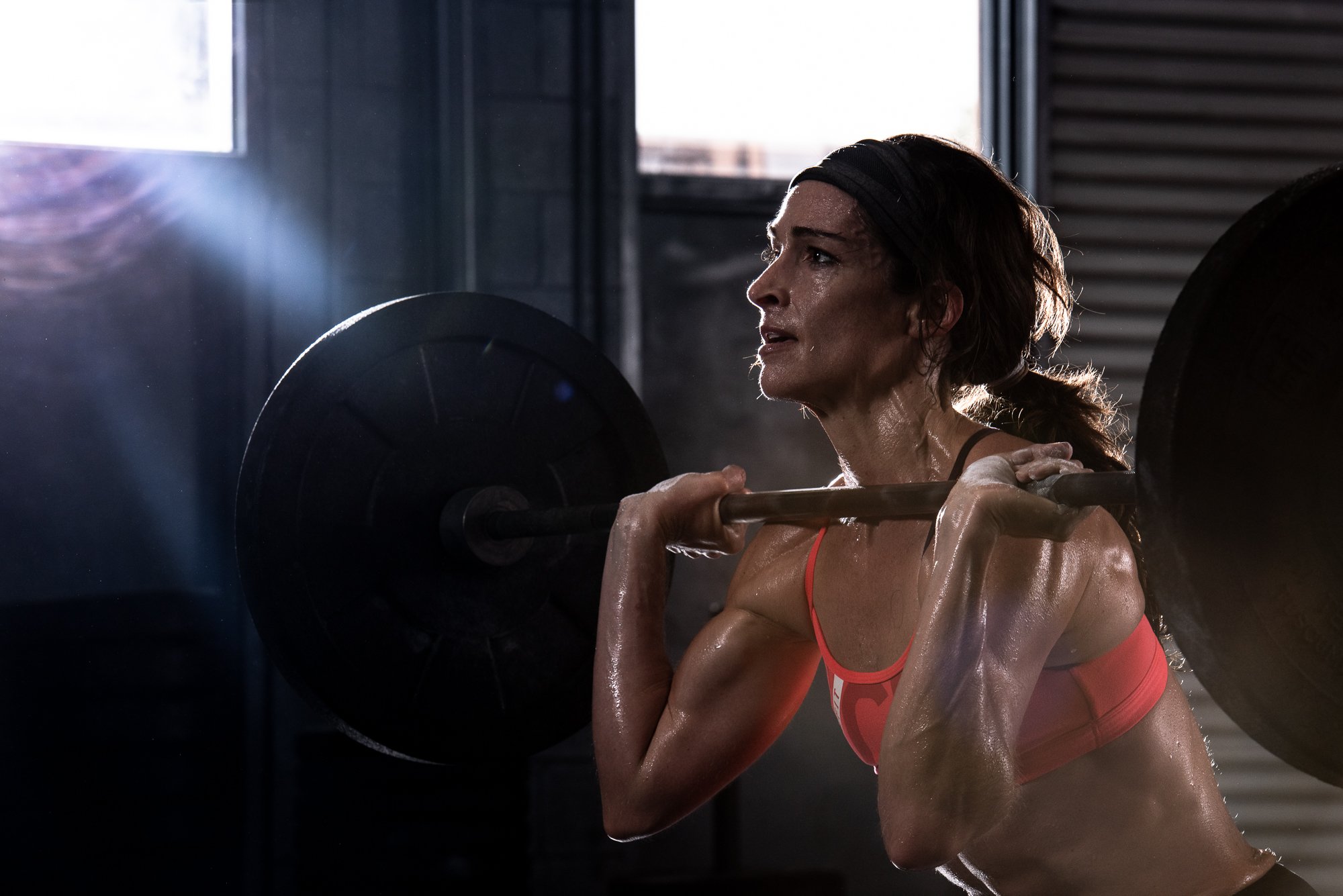 Female athlete performing barbell clean in gym with dramatic lighting and sweat during fitness photoshoot