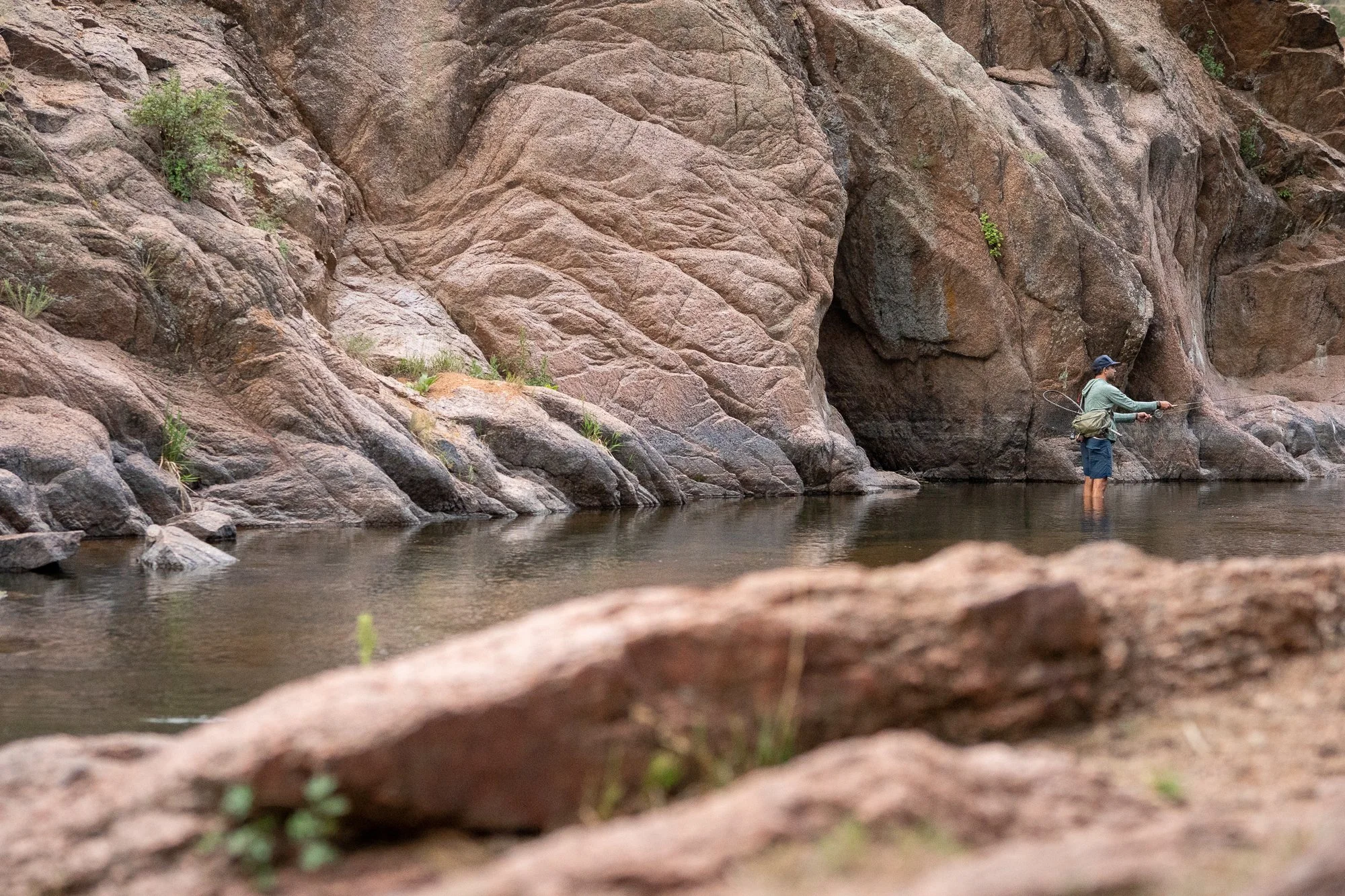 A fly angler stands in calm river water near a tall rock wall with a rod in hand