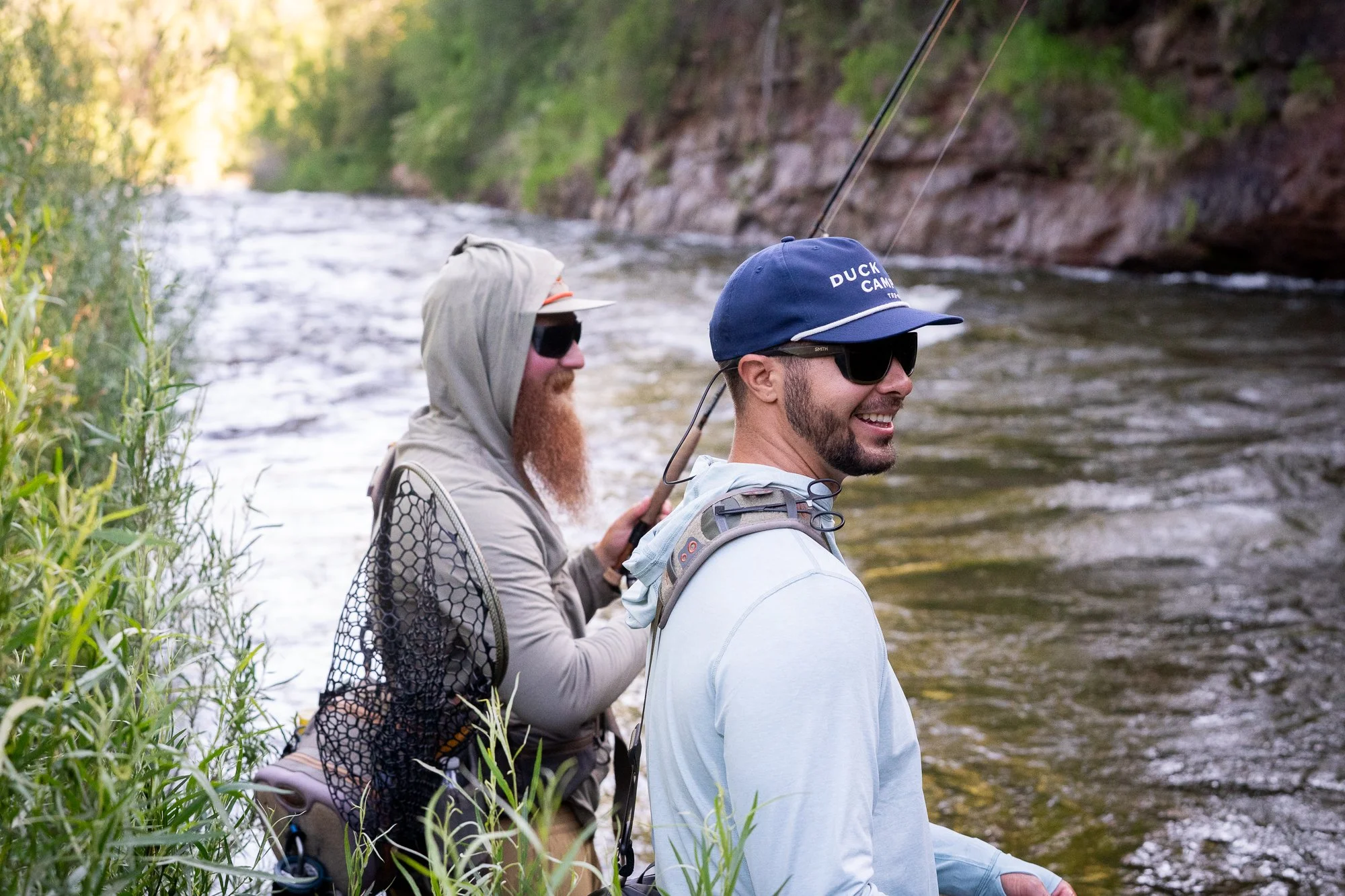 Two fly fishing anglers sit along the riverbank with rods and a landing net near the water