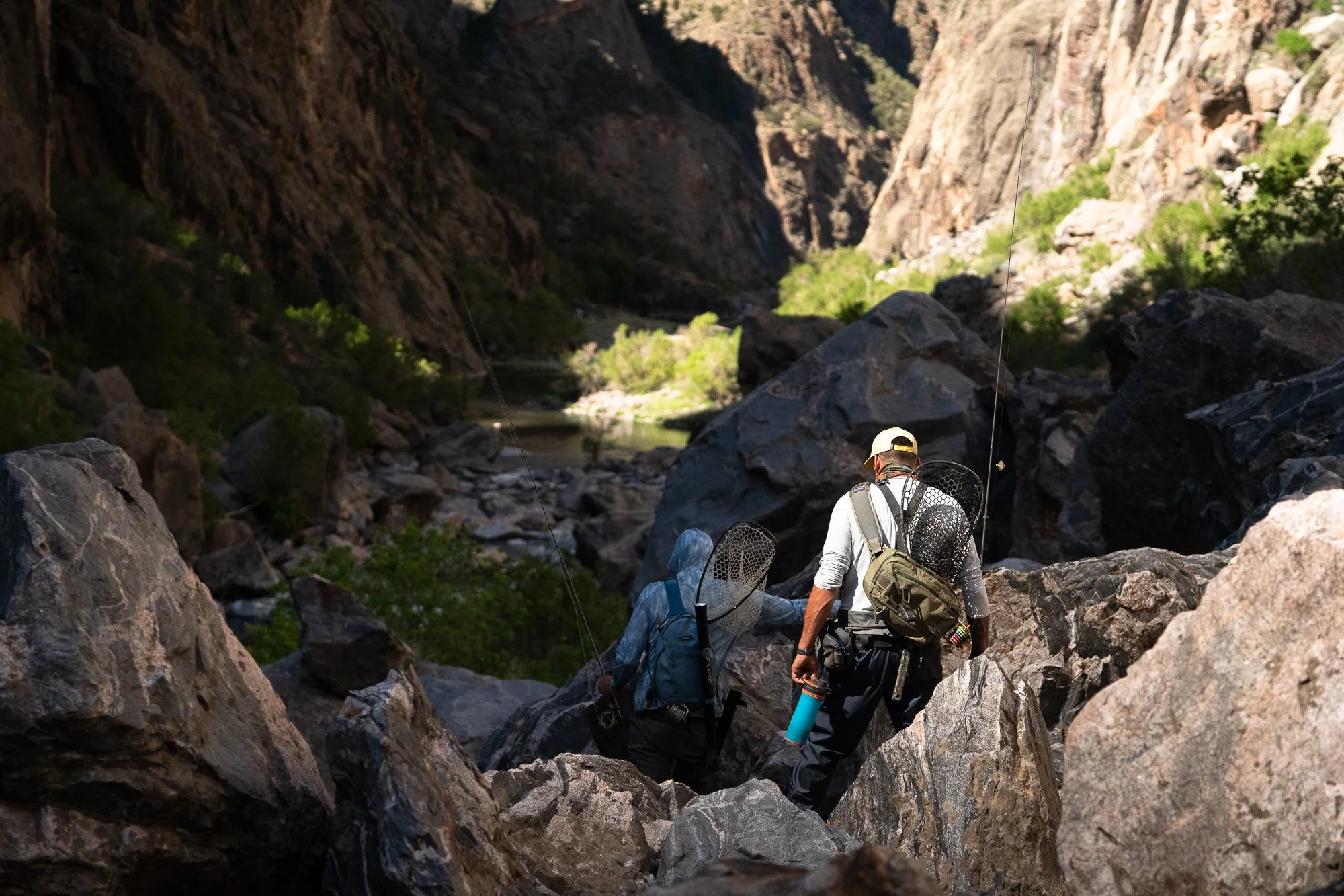 Two fly anglers climb through a rocky canyon with rods, packs, and landing nets