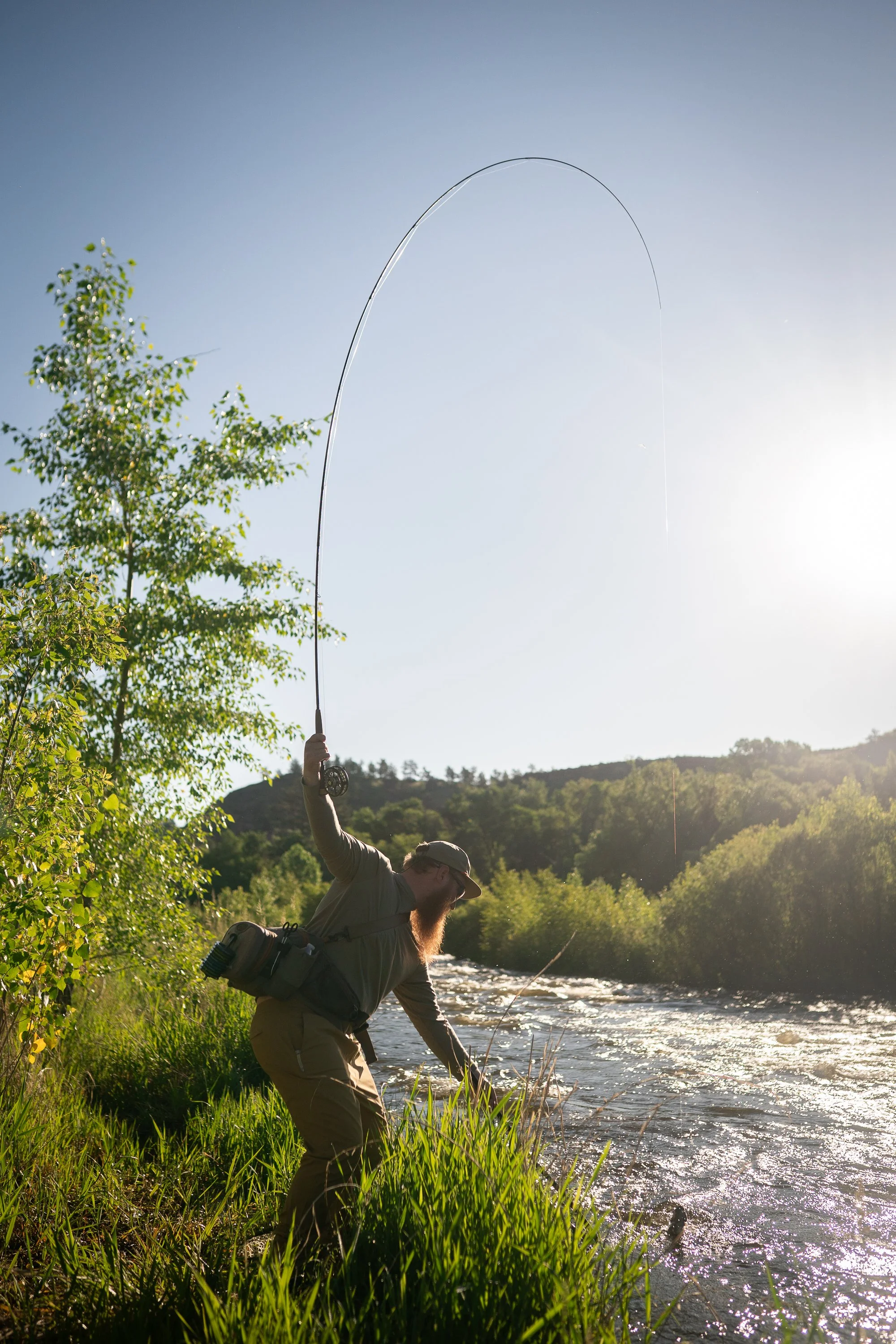 An angler lifts a bent fly rod along the riverbank with sunlight over the water