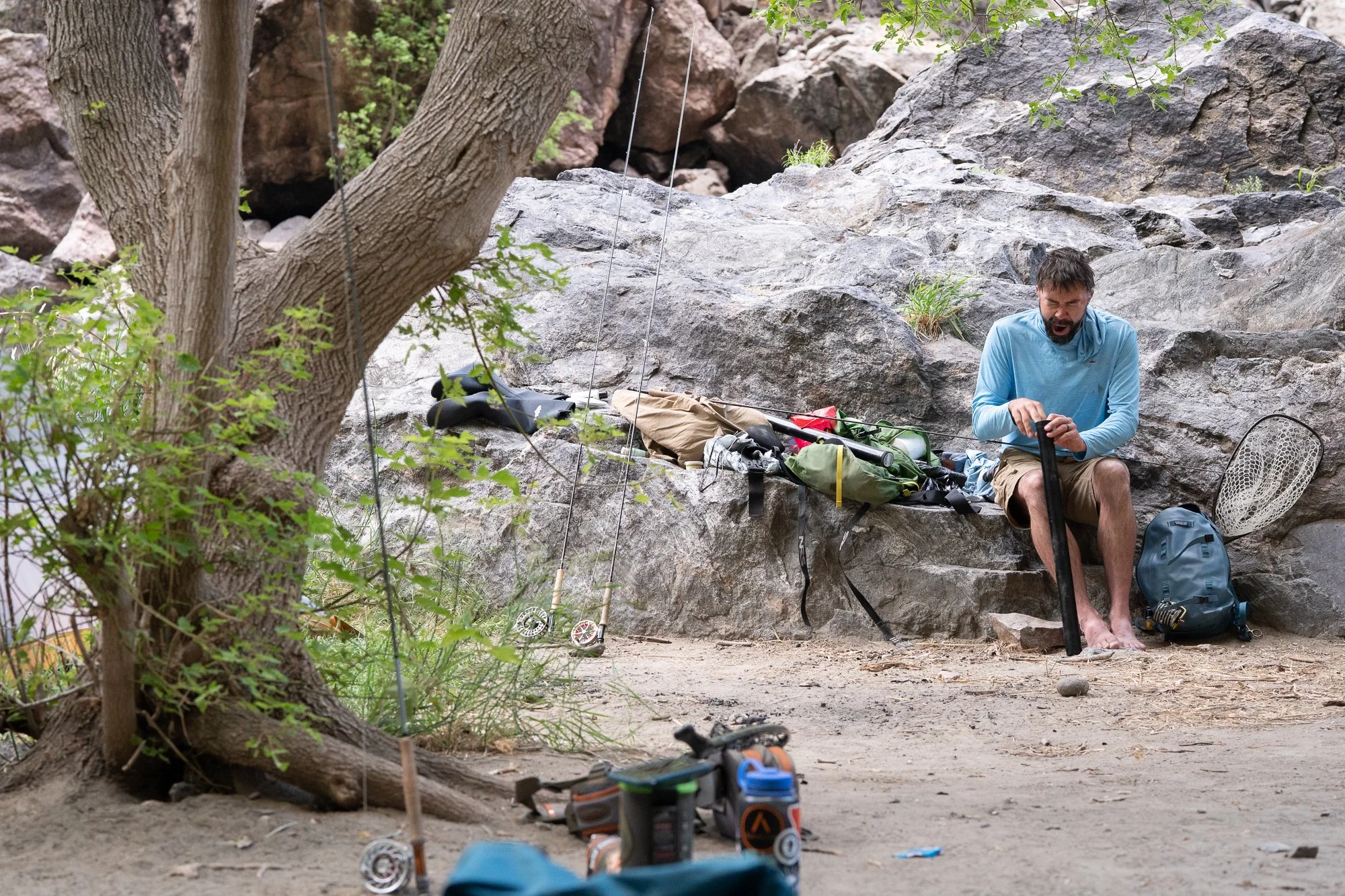 Fly fishing gear and rods set beside rocks while an angler prepares equipment near the river
