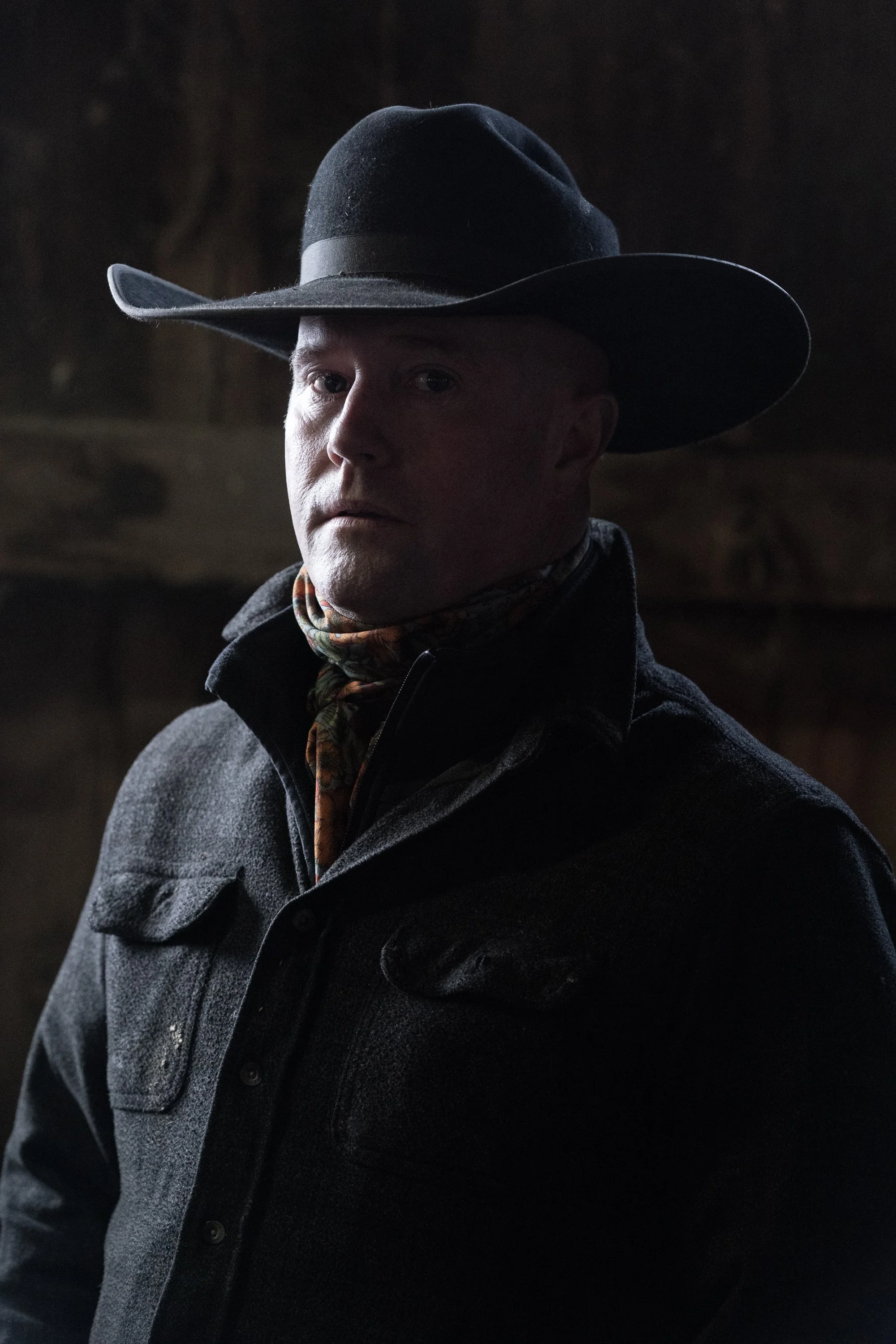 A cowboy in a black hat and dark coat stands for a low-light portrait inside a ranch building