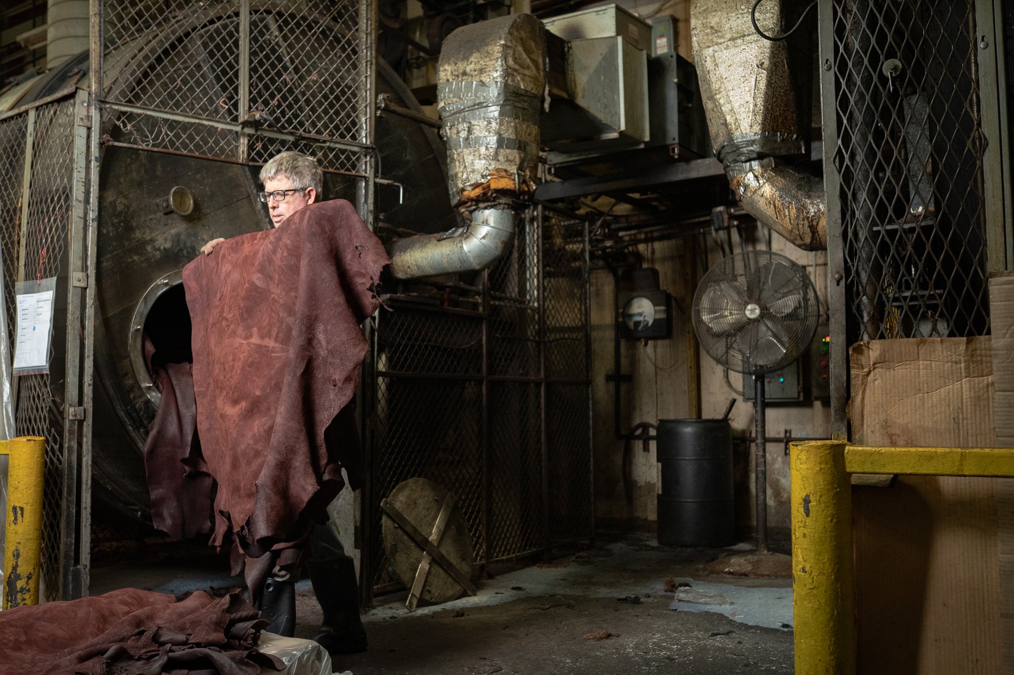 A folded leather hide is carried past metal machinery and stacked materials on the tannery floor