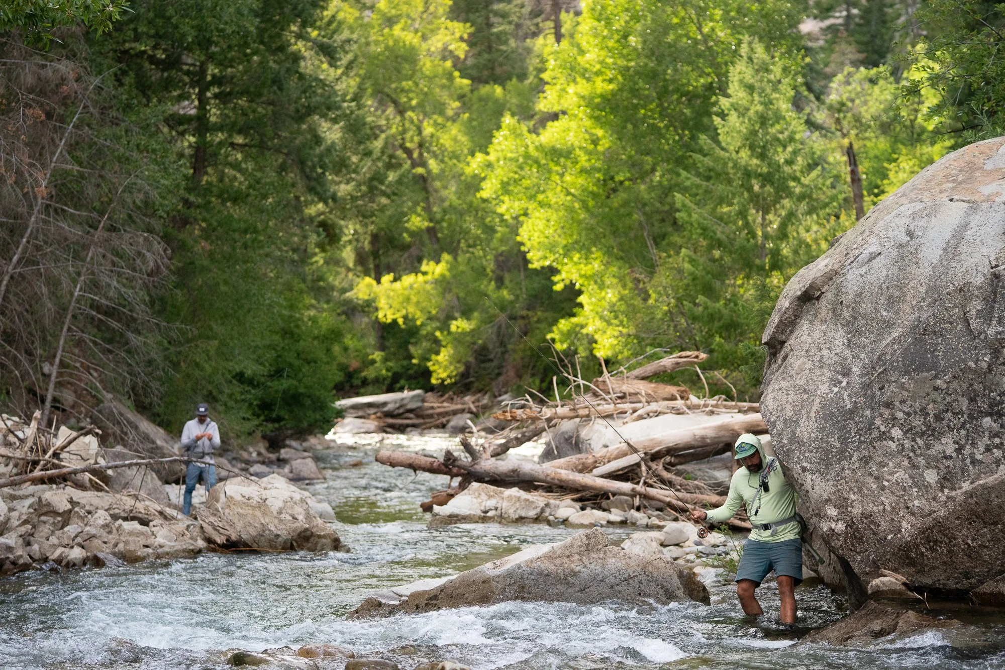 Two fly anglers work through a rocky river with trees and driftwood along the water