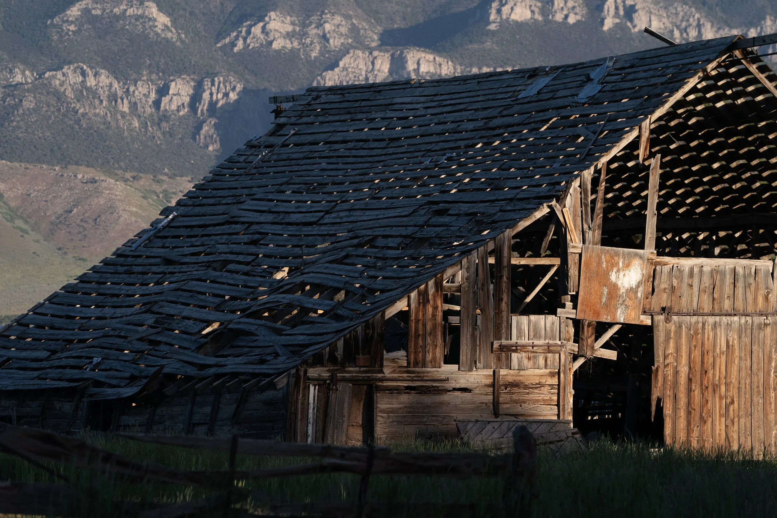 Weathered wooden ranch barn with a sagging, partially collapsed shingle roof and exposed timber framing against a rugged mountain landscape.