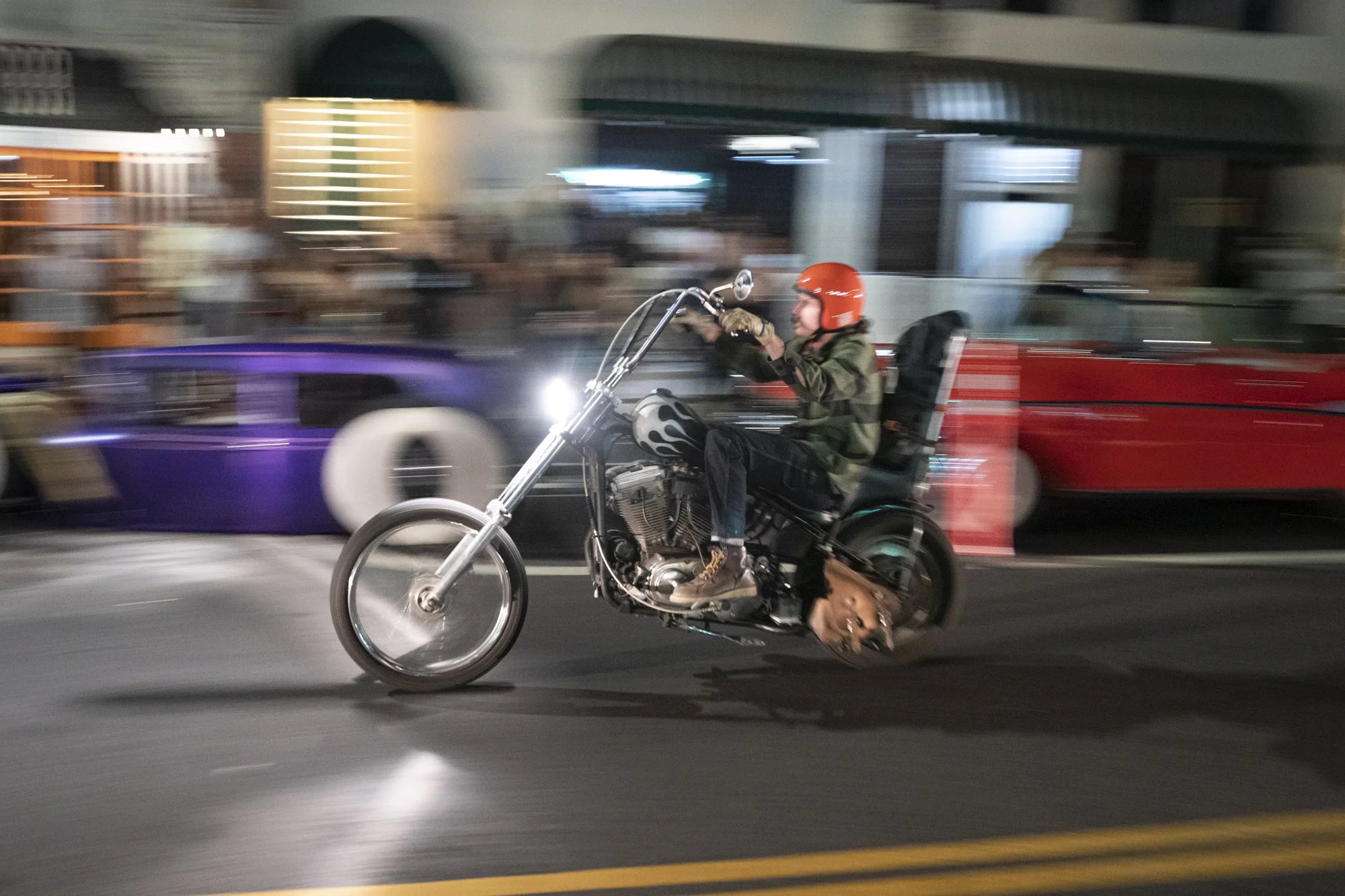 Chopper motorcycle with ape hanger handlebars and an orange helmet rider captured in motion blur on a Long Beach street during the Syndicate Barbershop anniversary night