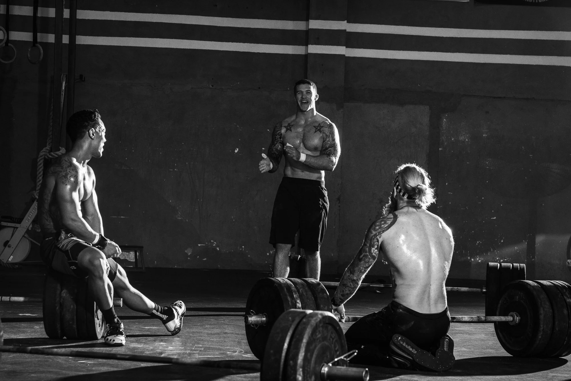 CrossFit athletes resting around loaded barbells during a workout in a gritty gym.