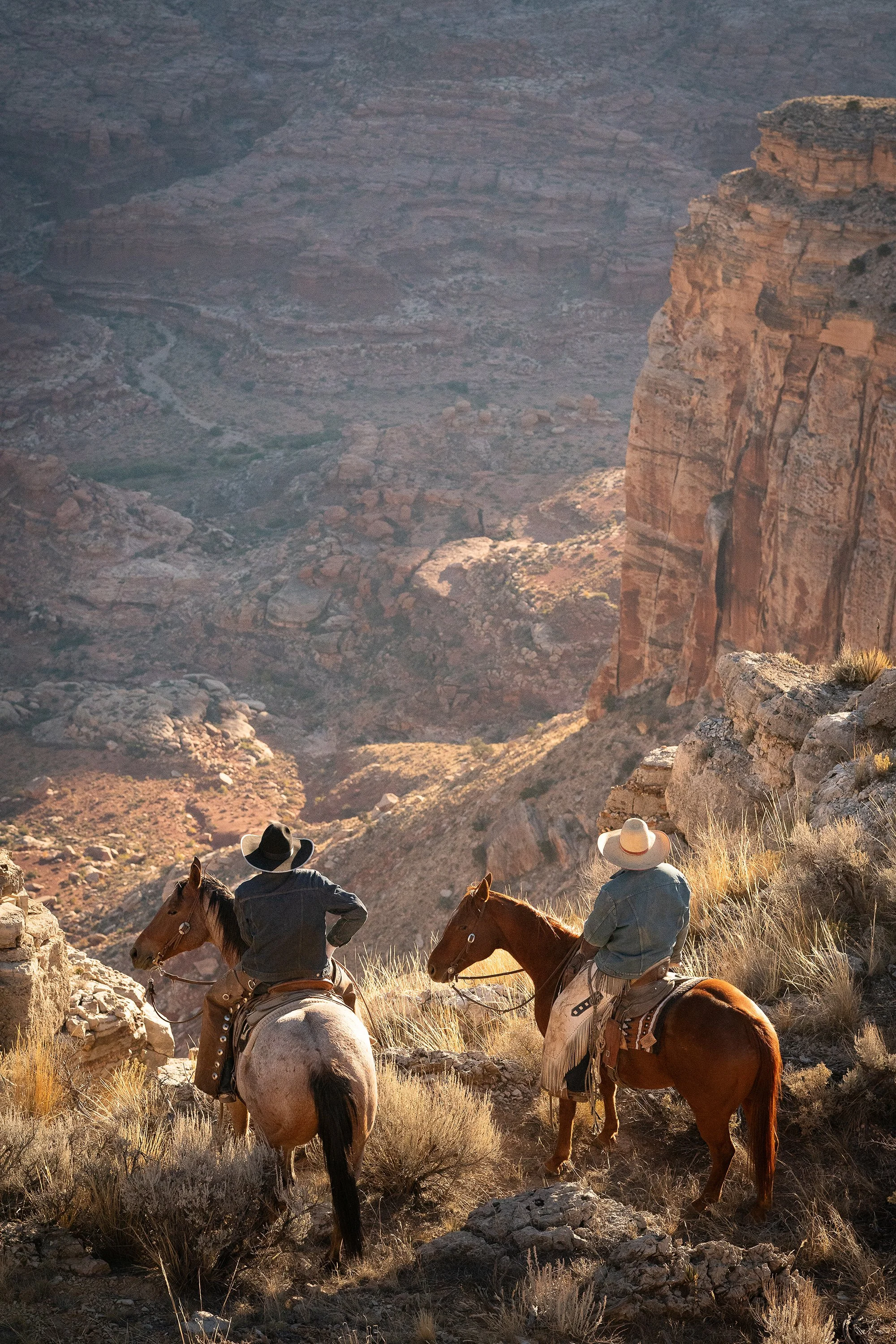 Two cowboys on horseback looking out over a deep red rock canyon in warm golden hour light from behind