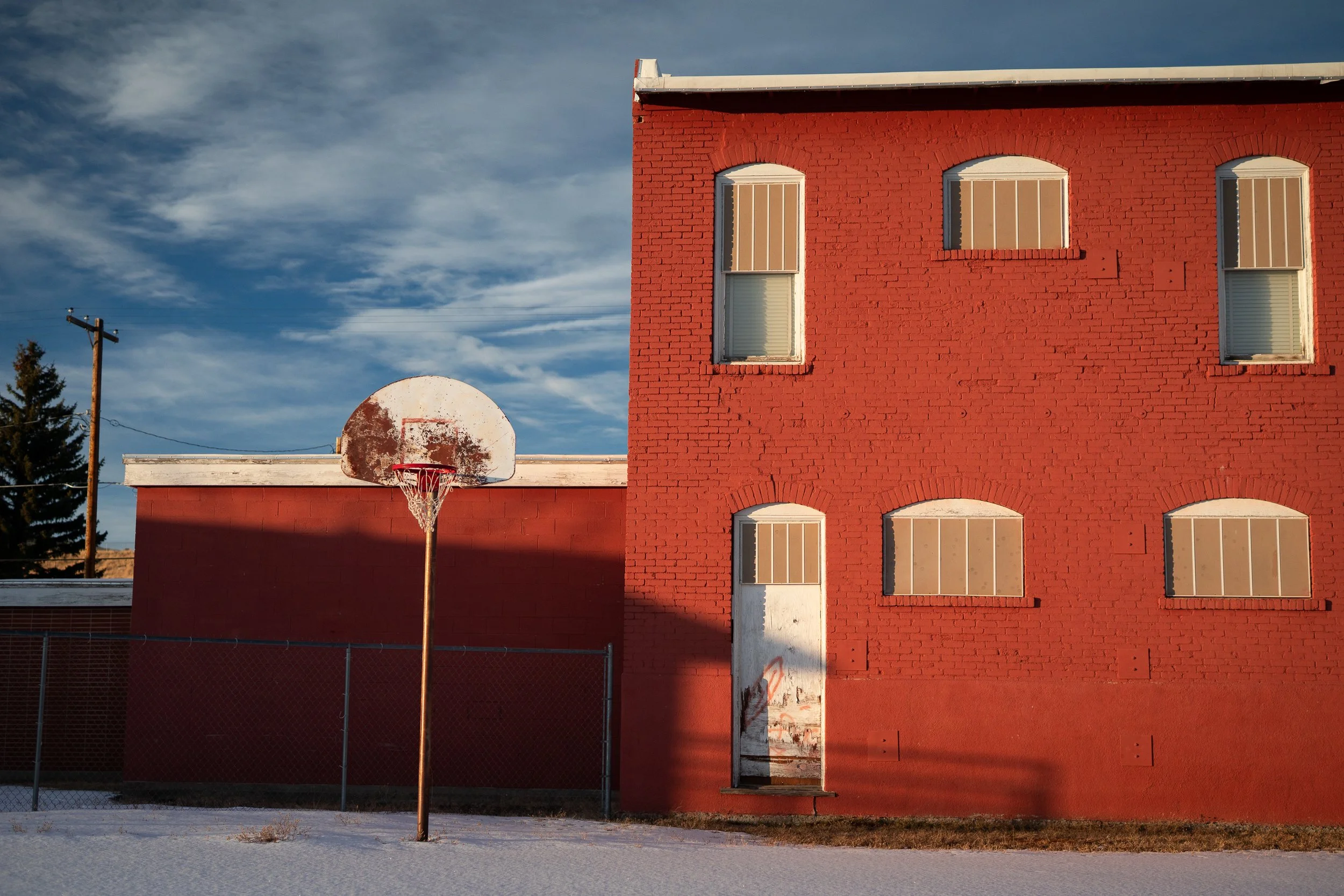 Weathered outdoor basketball hoop in front of a red brick building with snow on the ground.