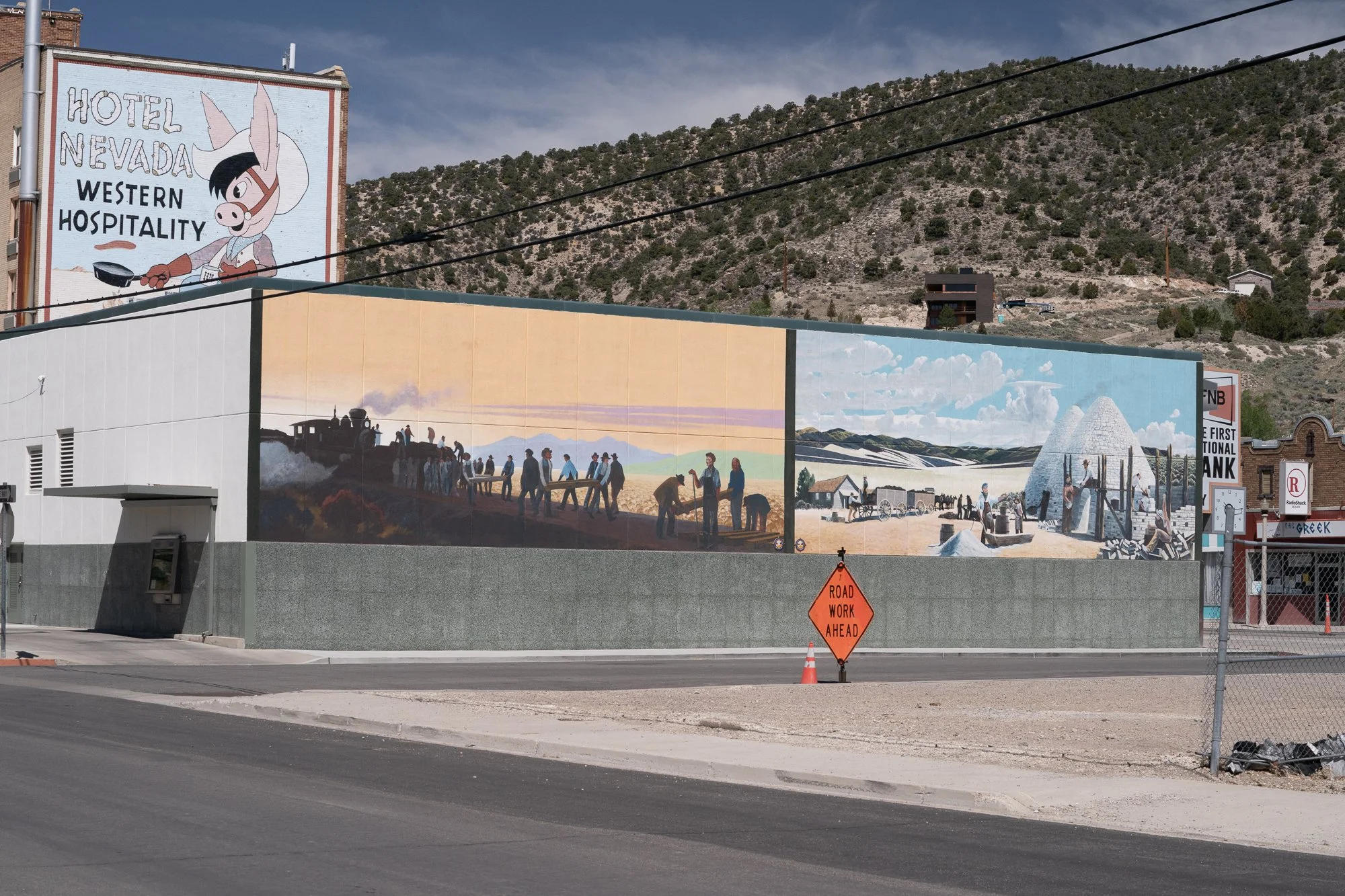 Downtown Ely, Nevada street corner with a large historic mural on a building wall, a Hotel Nevada Western Hospitality sign above, and a Road Work Ahead sign on the sidewalk.