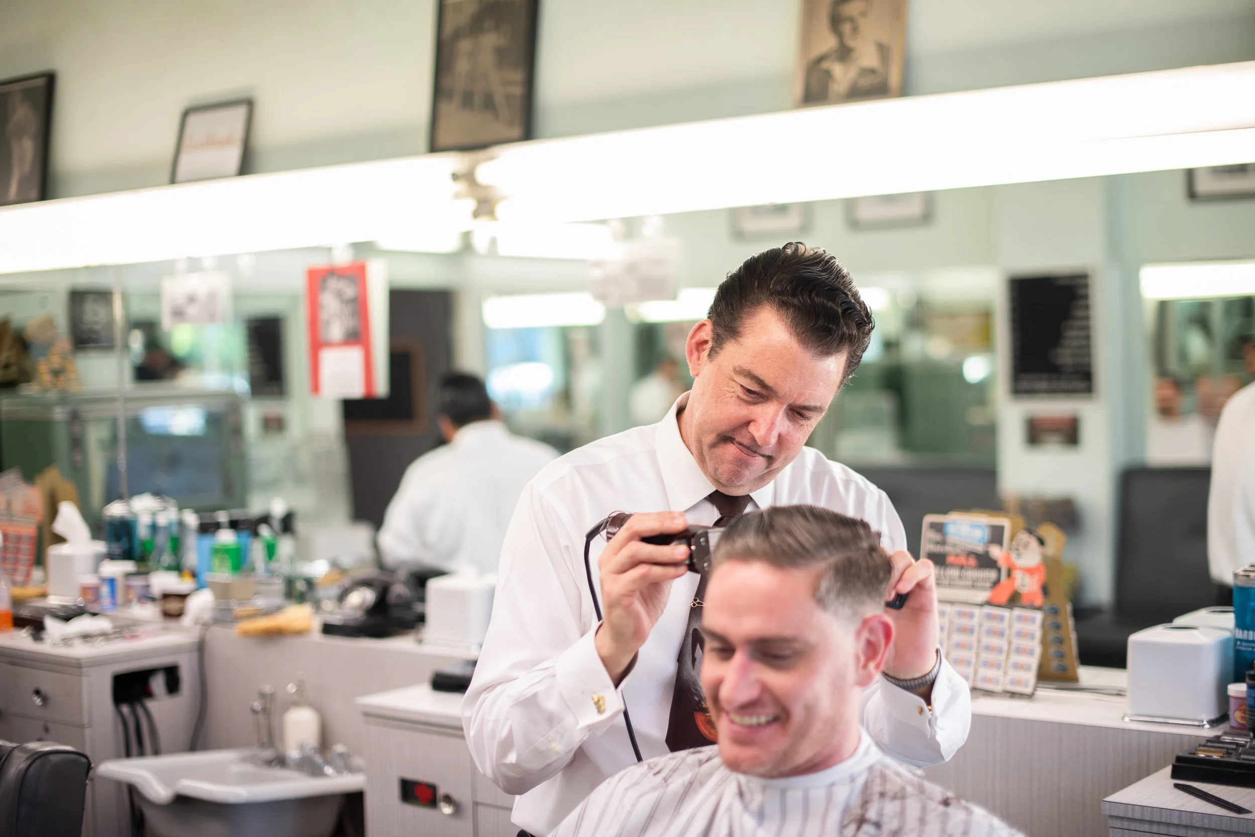 Barber in white shirt and dark tie smiling while using clippers on a laughing client's head at Sweeney Todd's Barbershop, mirrors and vintage wall decor visible behind them