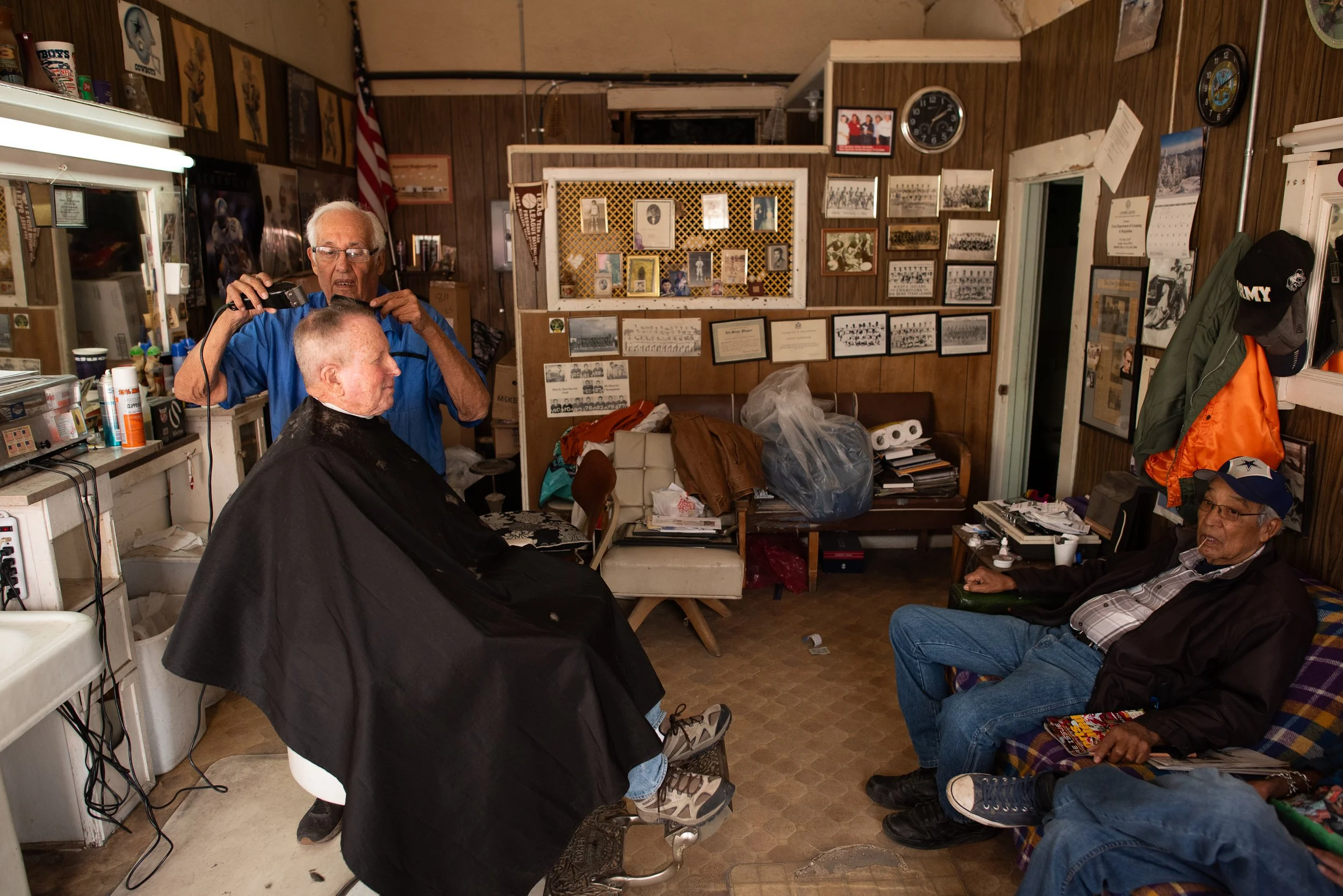 Customers sitting inside a wood-paneled barbershop in Marfa, Texas as a haircut continues.