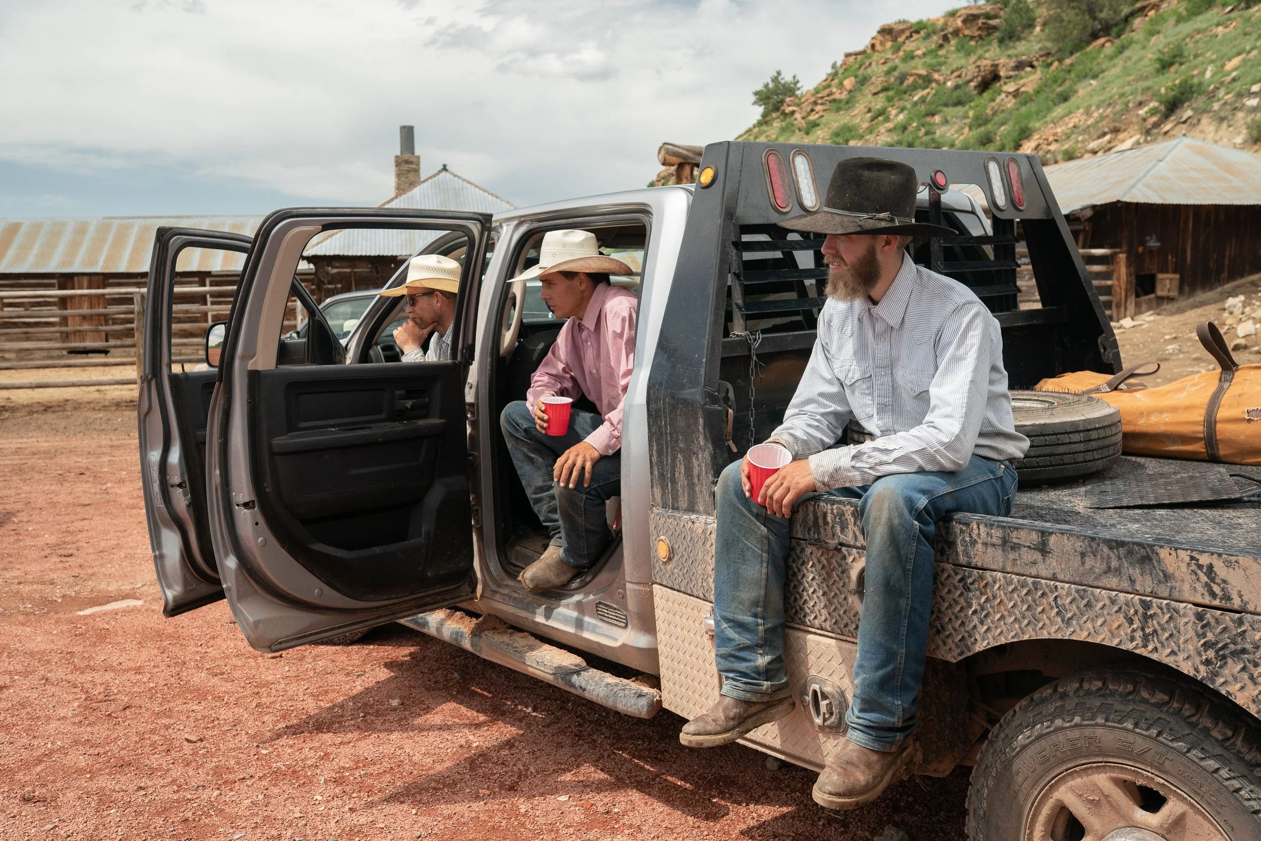 Cowboys sitting on and inside a pickup truck holding drinks while watching horse training on a Montana ranch