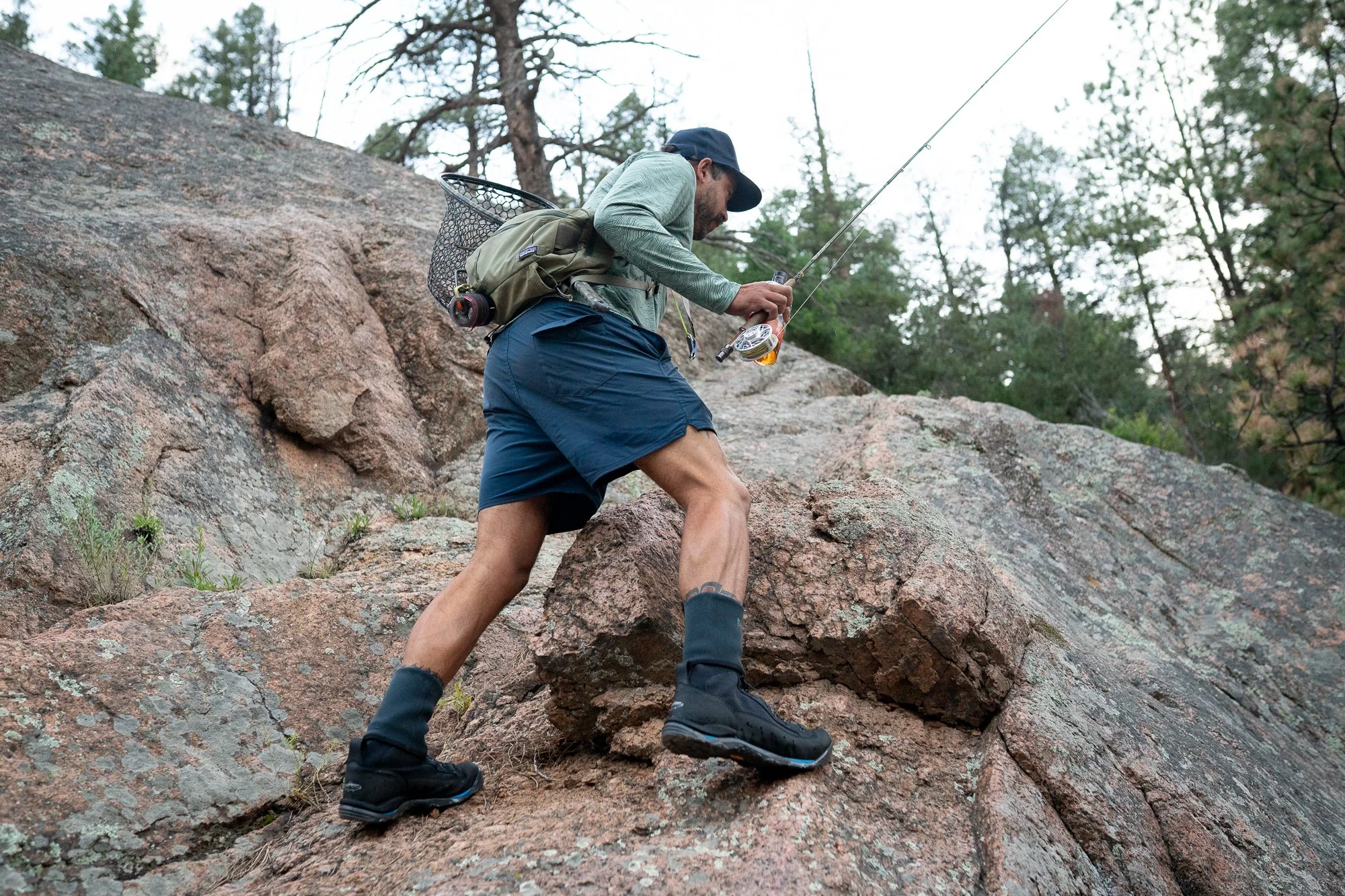 A fly angler climbs across large rocks while carrying a rod and landing net