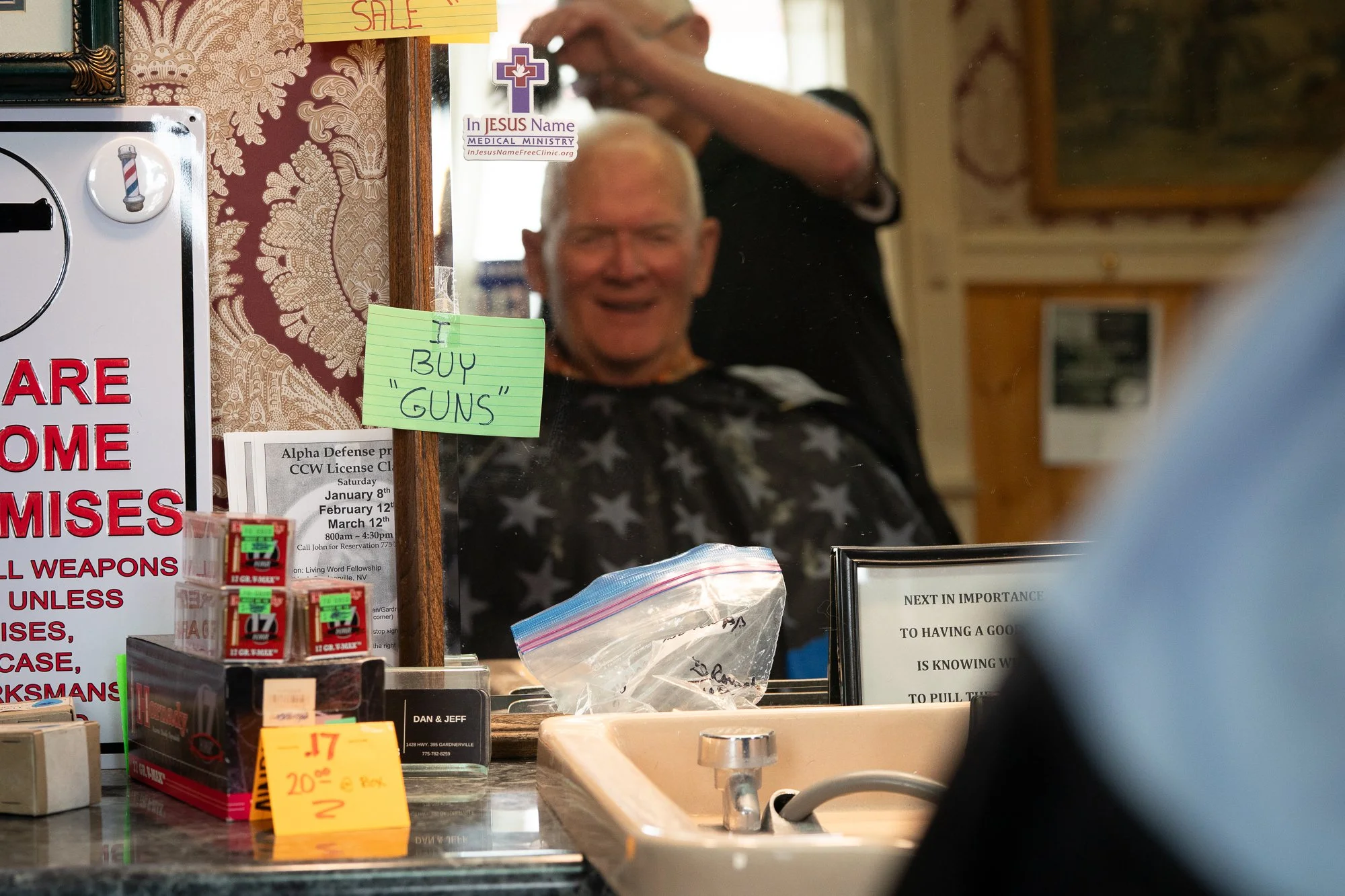 Nevada barbershop interior with a customer getting a haircut, handwritten and printed signs on the mirror including gun buying notice, ammunition boxes on the counter.