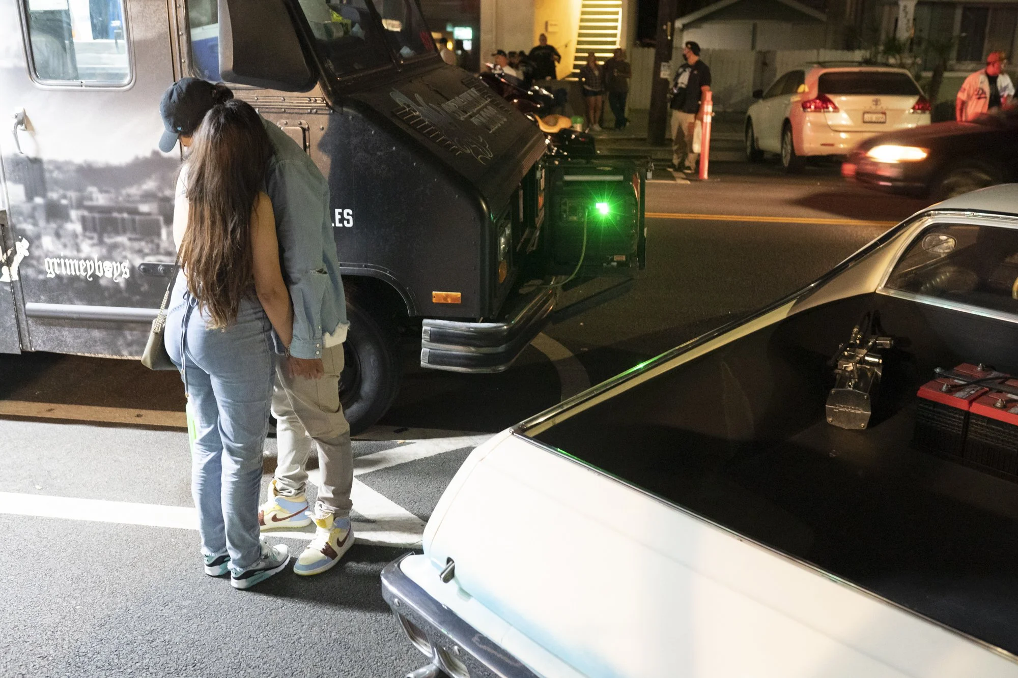Couple standing together looking into the open trunk of a white lowrider parked beside a black Grimey Boys truck in a Long Beach alley at night
