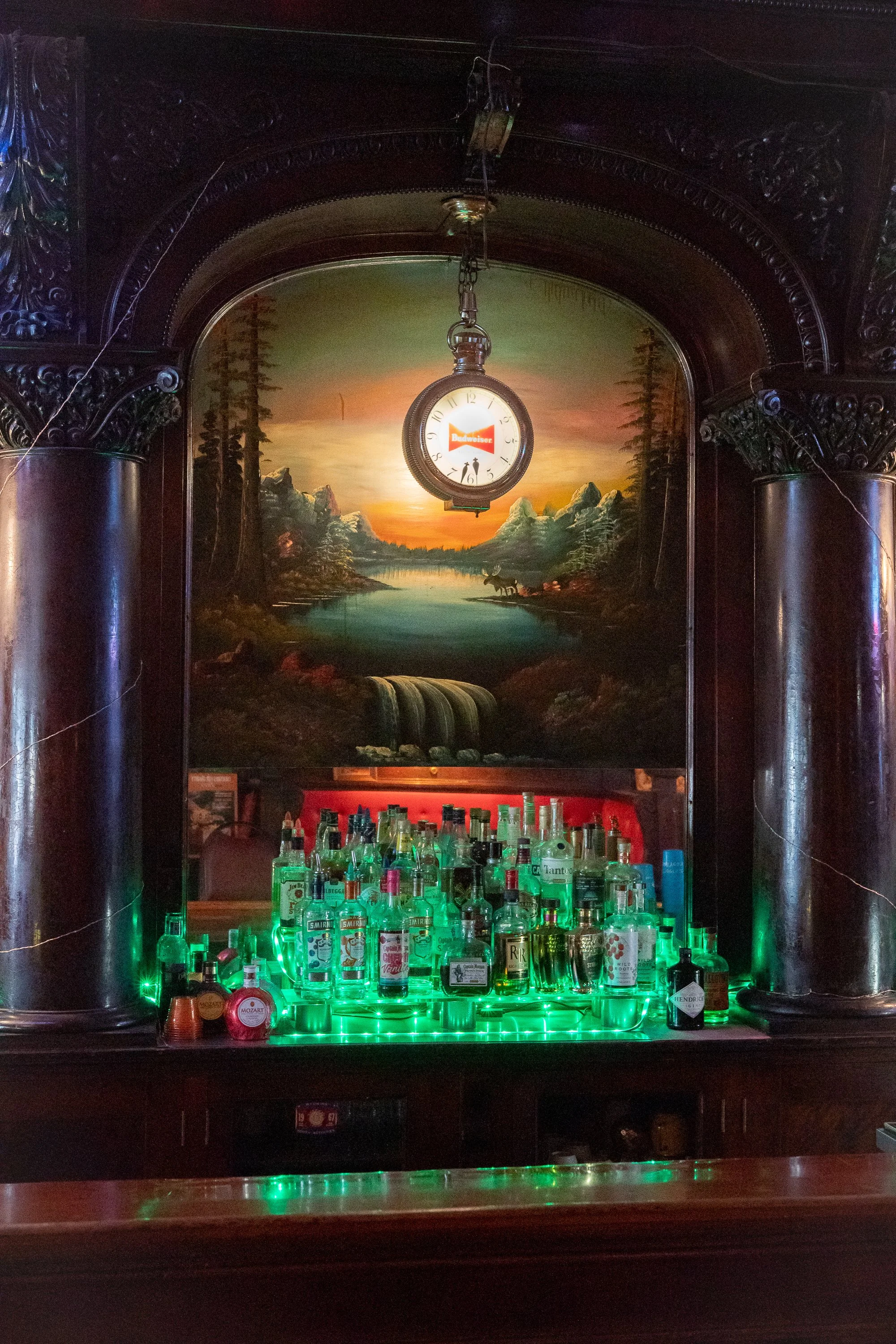 Liquor bottles illuminated by green lights beneath a hanging Budweiser clock and a painted landscape inside the College Inn in Douglas, Wyoming.