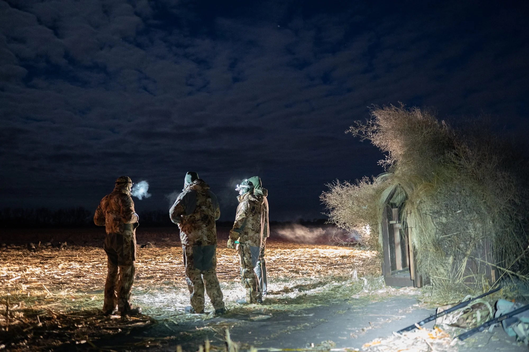 Duck hunters stand beside a camouflaged field blind before dawn