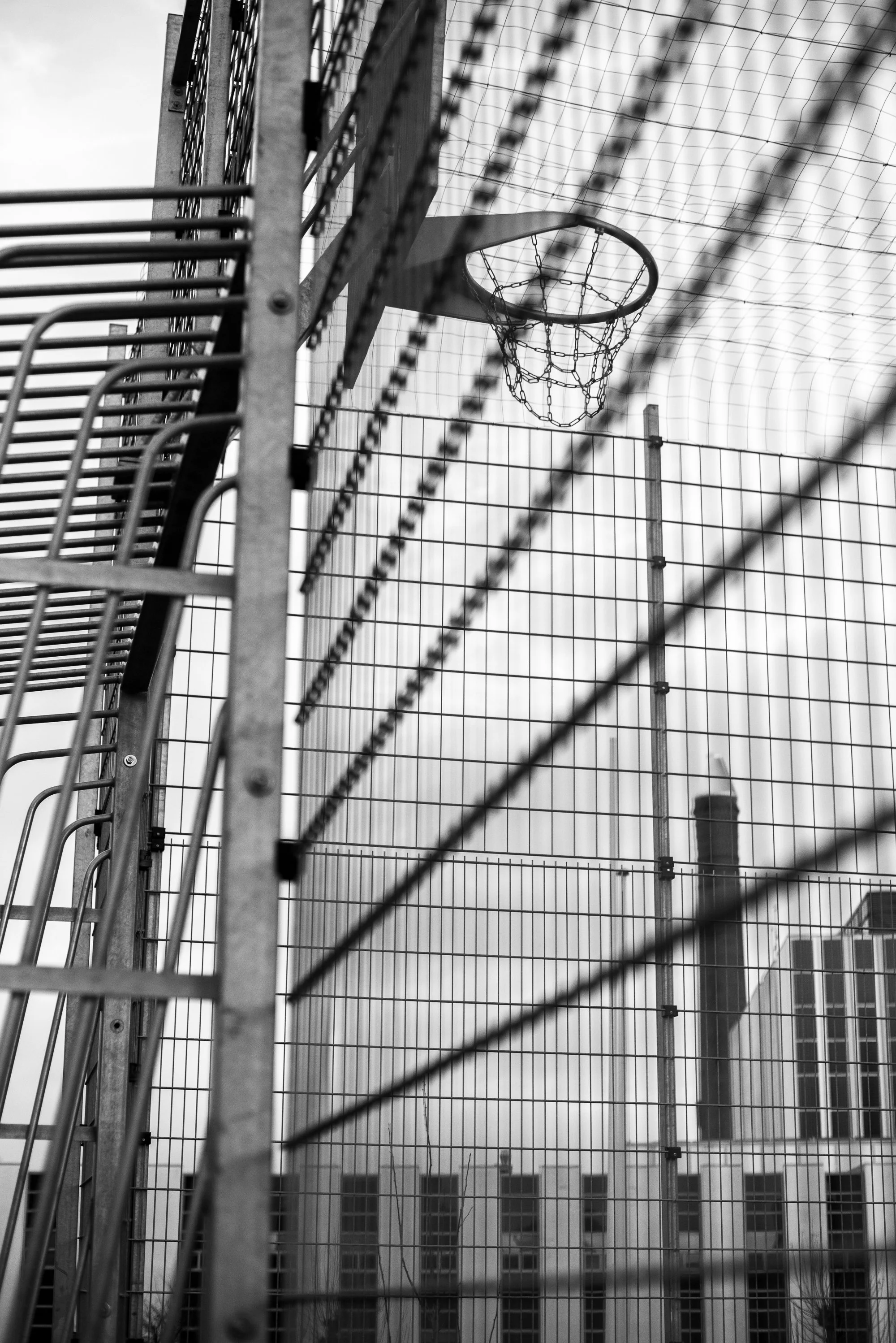 Black and white photograph of a basketball hoop viewed through fencing on an outdoor European court.