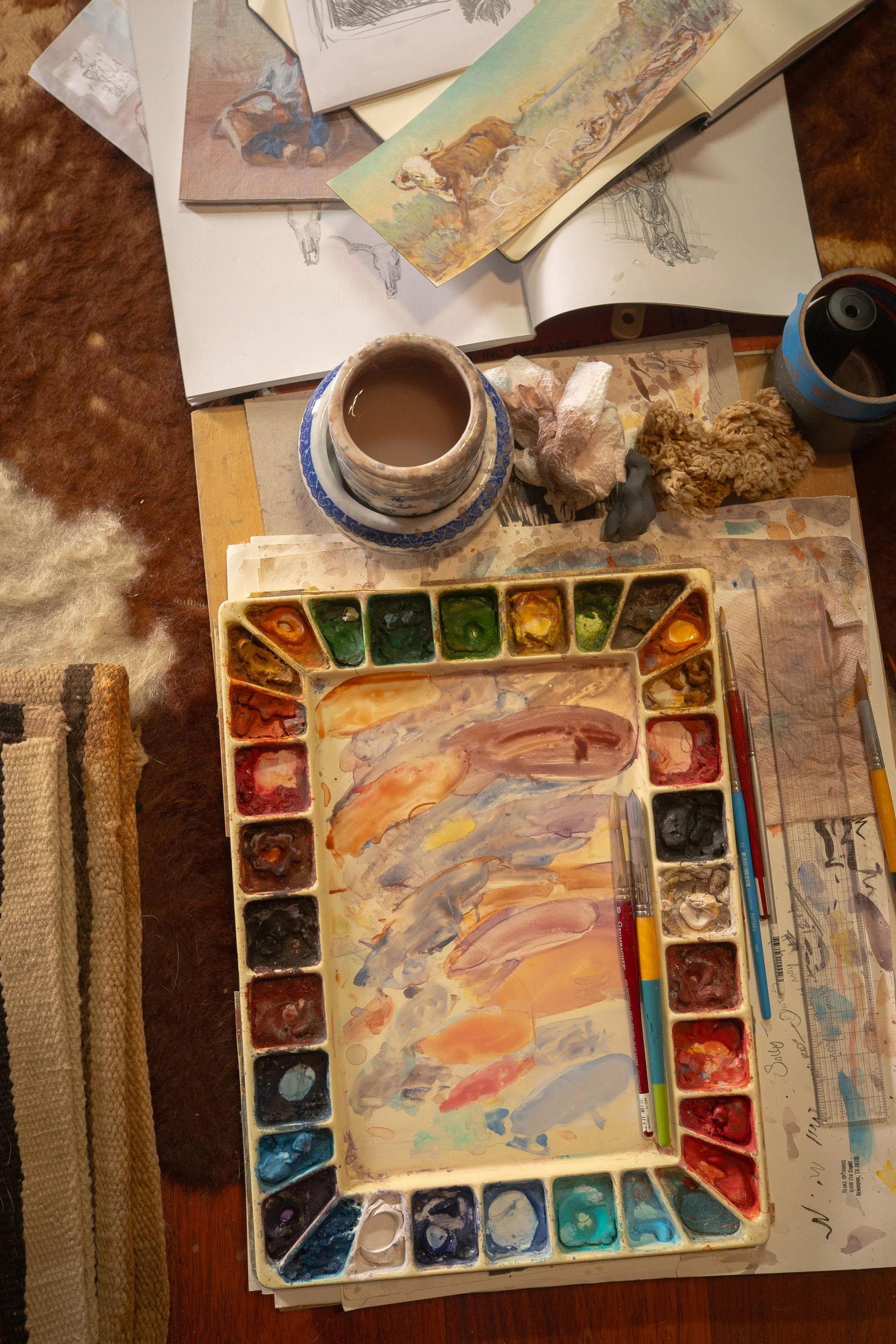 Overhead view of watercolor palette, brushes, and sketches inside Western artist Teal Blake’s studio