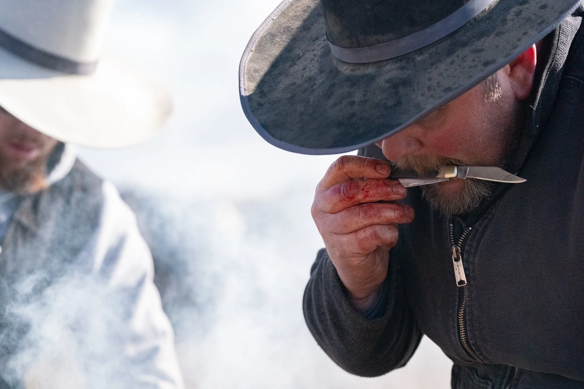 A cowboy holds a small knife close to his mouth during cattle work, with smoke and another cowboy in the background